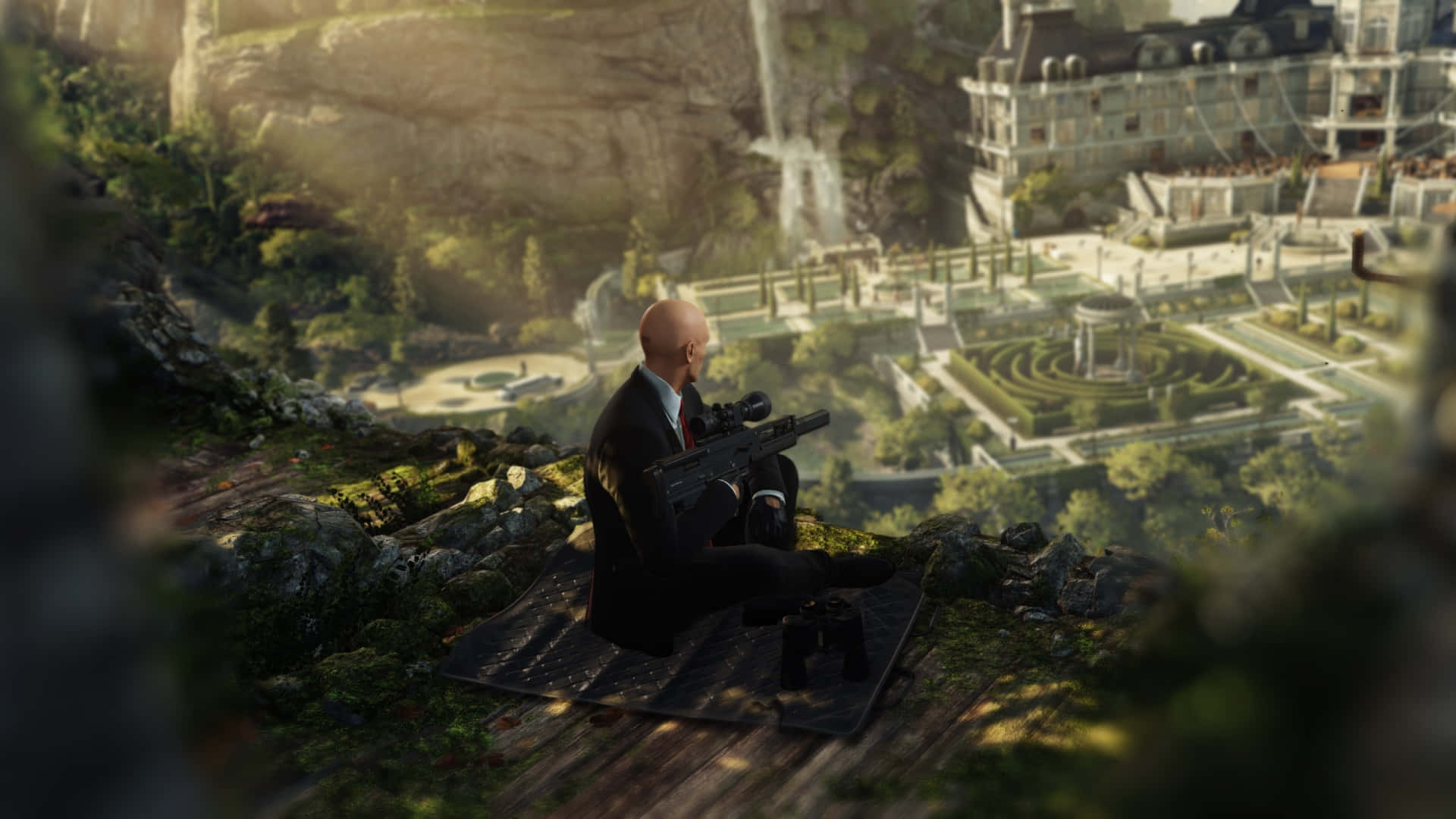 《Hitman 2》特工執行任務 壁紙 — 一個光頭男子穿著西裝坐在懸崖頂上，俯瞰著一個宏偉的莊園，有瀑布和修剪整齊的花園沐浴在金色的陽光下。