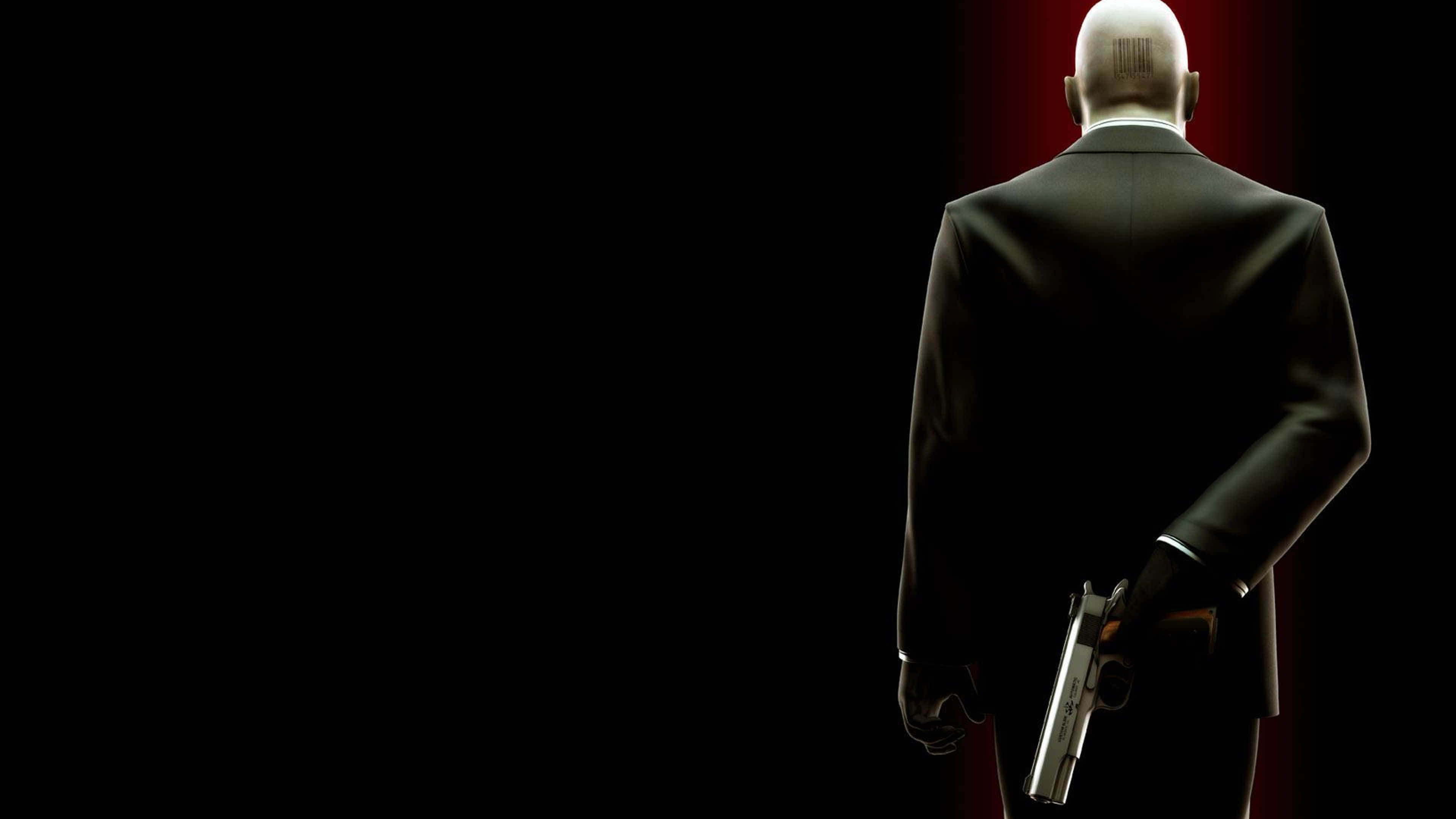 Hitman背後持槍桌面壁紙 壁紙 — 一個光頭男人穿著黑色西裝，背對觀眾，在黑暗背景中手持銀色手槍，背景有紅色燈光。