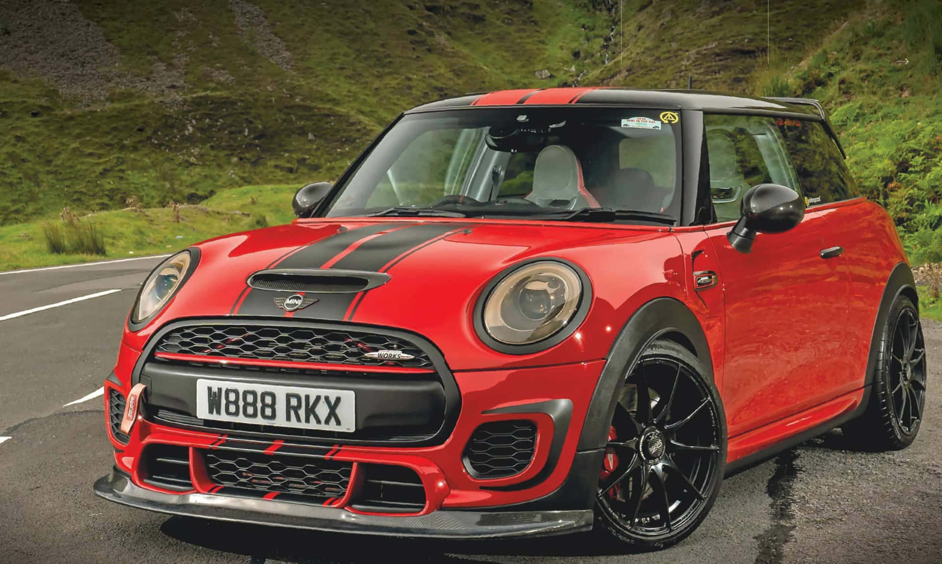  壁紙 — 一輛帶有黑色賽車條紋的鮮紅色MINI John Cooper Works在蜿蜒的山路上行駛，周圍是鬱鬱蔥蔥的綠色山丘。