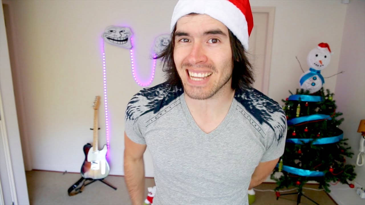 HolaSoyGerman 度過聖誕節 壁紙 — 一個戴著聖誕帽的男人在一個裝飾著亮燈聖誕樹和發光巨魔臉項鍊的房間裡咧嘴大笑。