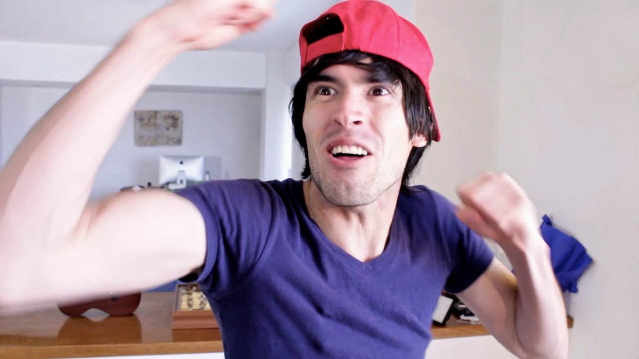 HolaSoyGerman 戴反帽 壁紙 — 一個戴紅色帽同紫色衫嘅男人喺明亮嘅室內房間入面，興奮咁屈起雙臂。