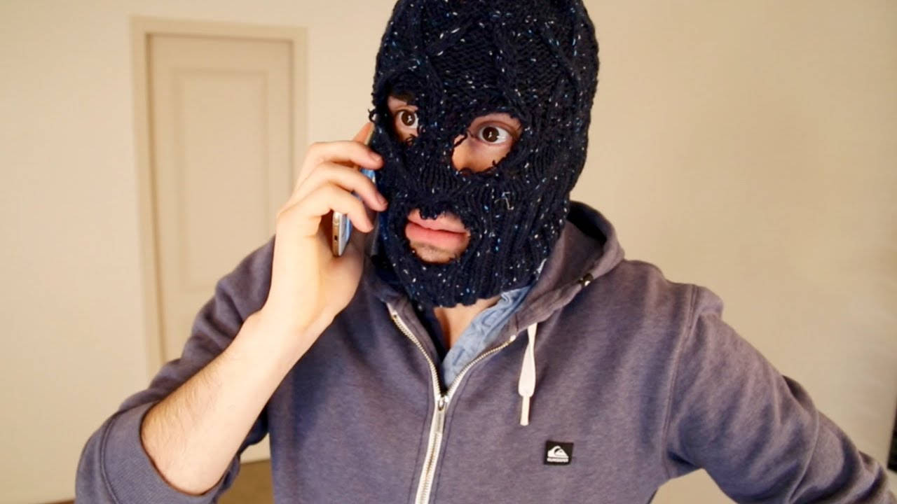 HolaSoyGerman 戴黑色口罩 壁紙 — 一個戴著深色針織巴拉克拉瓦帽的人，在室內打電話，身後是樸素的牆壁和門。