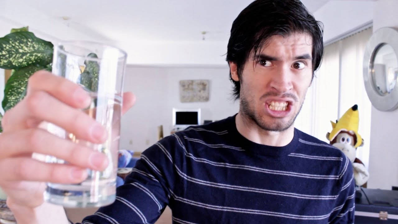 HolaSoyGerman 手持水杯怒視 壁紙 — 一名深色頭髮、身穿條紋襯衫的男子在室內舉起一杯水，做出誇張的苦臉。