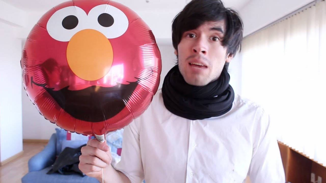 HolaSoyGerman 手持艾摩氣球 壁紙 — 一位身穿白恤衫的年輕男子在室內窗邊拿著一個大型紅色Elmo氣球。