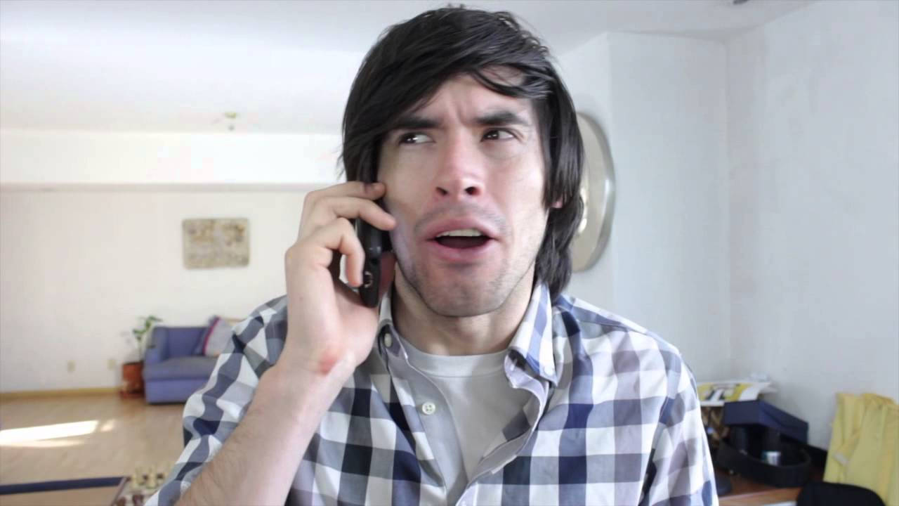 HolaSoyGerman 打電話搞笑劇 壁紙 — 一個穿著格仔恤衫的男人在明亮的客廳裡，拿著電話貼著耳朵，看起來很震驚。