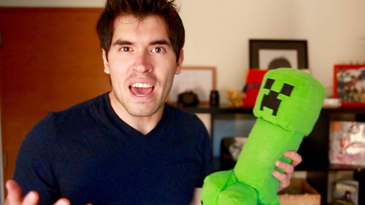 HolaSoyGerman 拿綠色毛絨玩具 壁紙 — 一個表情興奮的男人在室內拿著一個大型綠色Minecraft Creeper毛公仔。