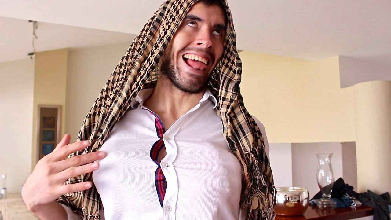 HolaSoyGerman 搞笑鬼臉 壁紙 — 一位留鬍鬚的男子在室內開懷大笑，同時頑皮地將一條格紋圍巾披在頭上。