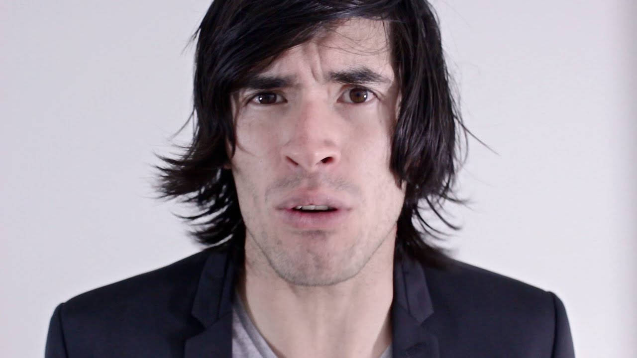 HolaSoyGerman 混淆表情 壁紙 — 一個頭髮深色凌亂、身穿黑色西裝外套的男人，在純白背景前睜大眼睛、微張嘴巴，強烈地凝視著。