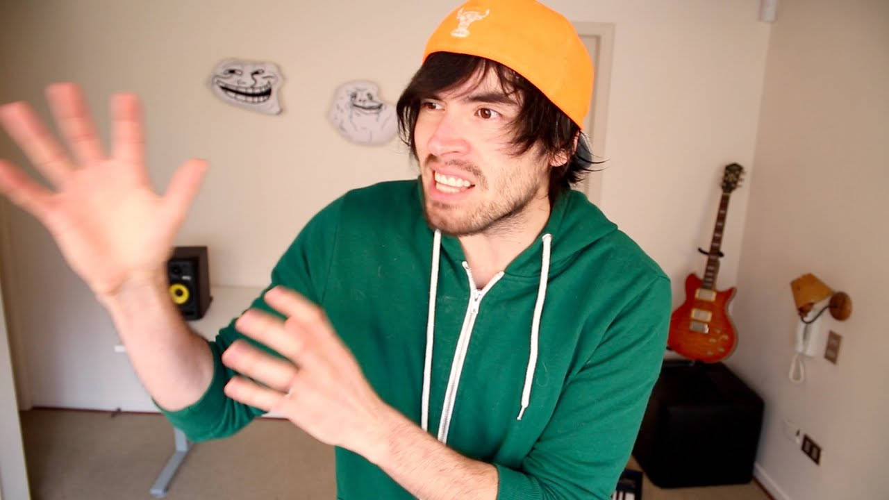 HolaSoyGerman 綠色連帽衫 壁紙 — 一名穿著綠色連帽衫和橙色帽子的男子在一個有紅色結他和 Troll Face 海報的房間裡生動地做手勢。