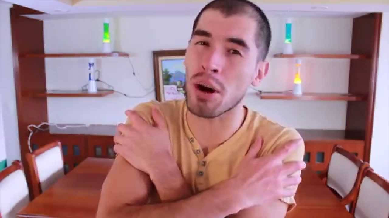 HolaSoyGerman 自抱自己 壁紙 — 一名身穿黃色襯衫的男子在一個有木製家具和發光熔岩燈的飯廳中雙臂交叉放在胸前。