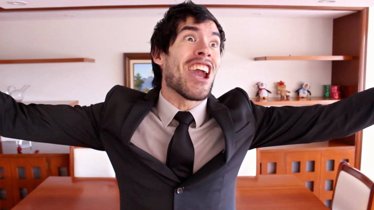 HolaSoyGerman 西裝領帶 壁紙 — 一名身穿黑色西裝的男子站在一個有木製家具的房間裡，雙臂張開，表情狂喜。