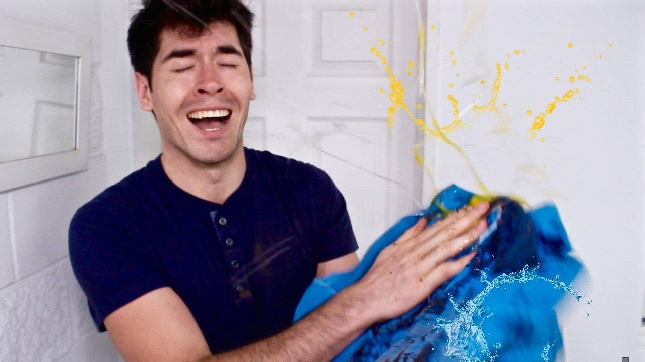HolaSoyGerman 輕撫藍布 壁紙 — 一名男子開心大笑，同時擠壓一塊藍色布料，令黃色和藍色液體戲劇性地濺到白色牆壁上。