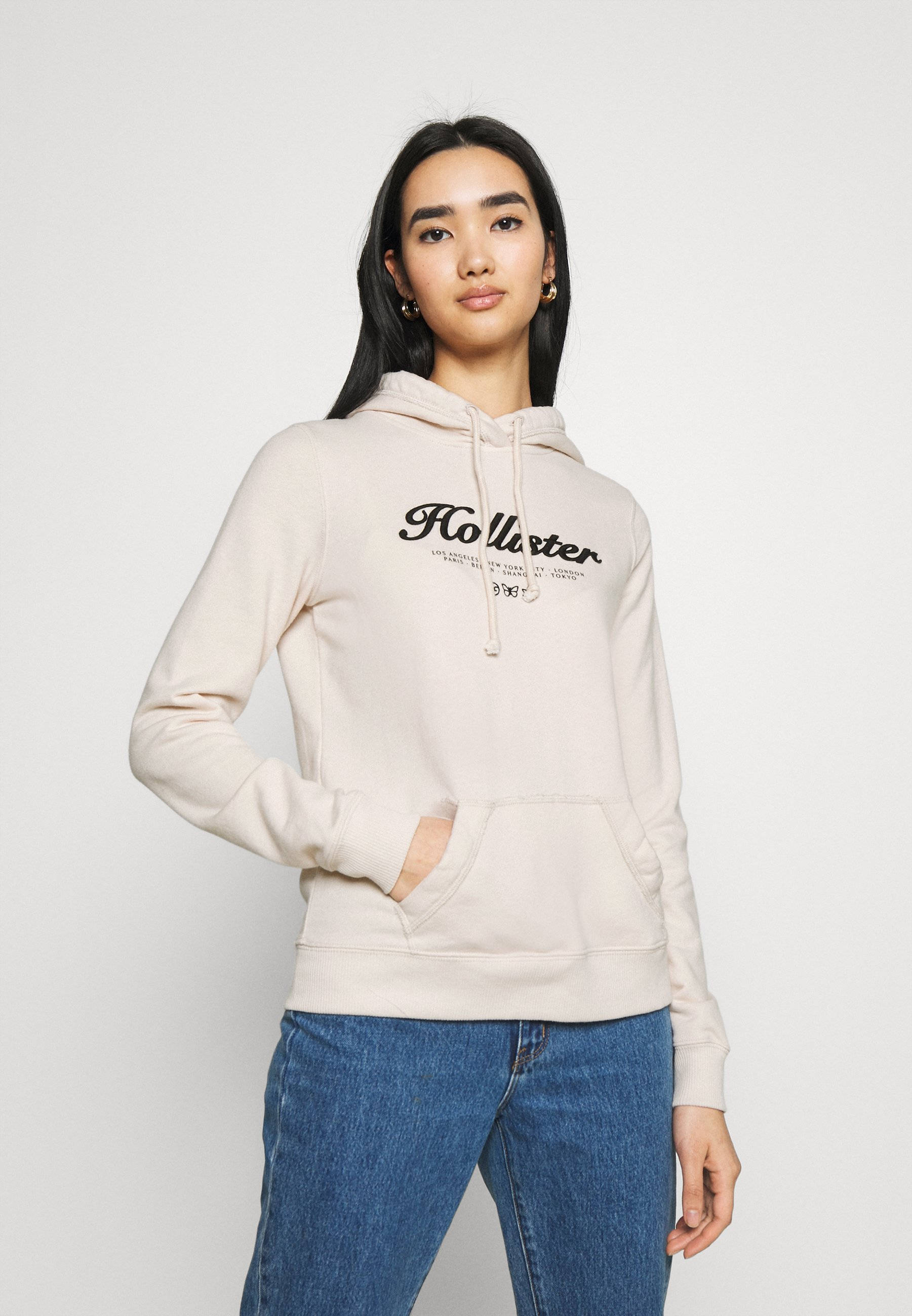 米白色 Hollister 連帽衛衣 壁紙 — 一位穿著米色Hollister連帽衫和藍色牛仔褲的女性自信地站在純白色背景前。