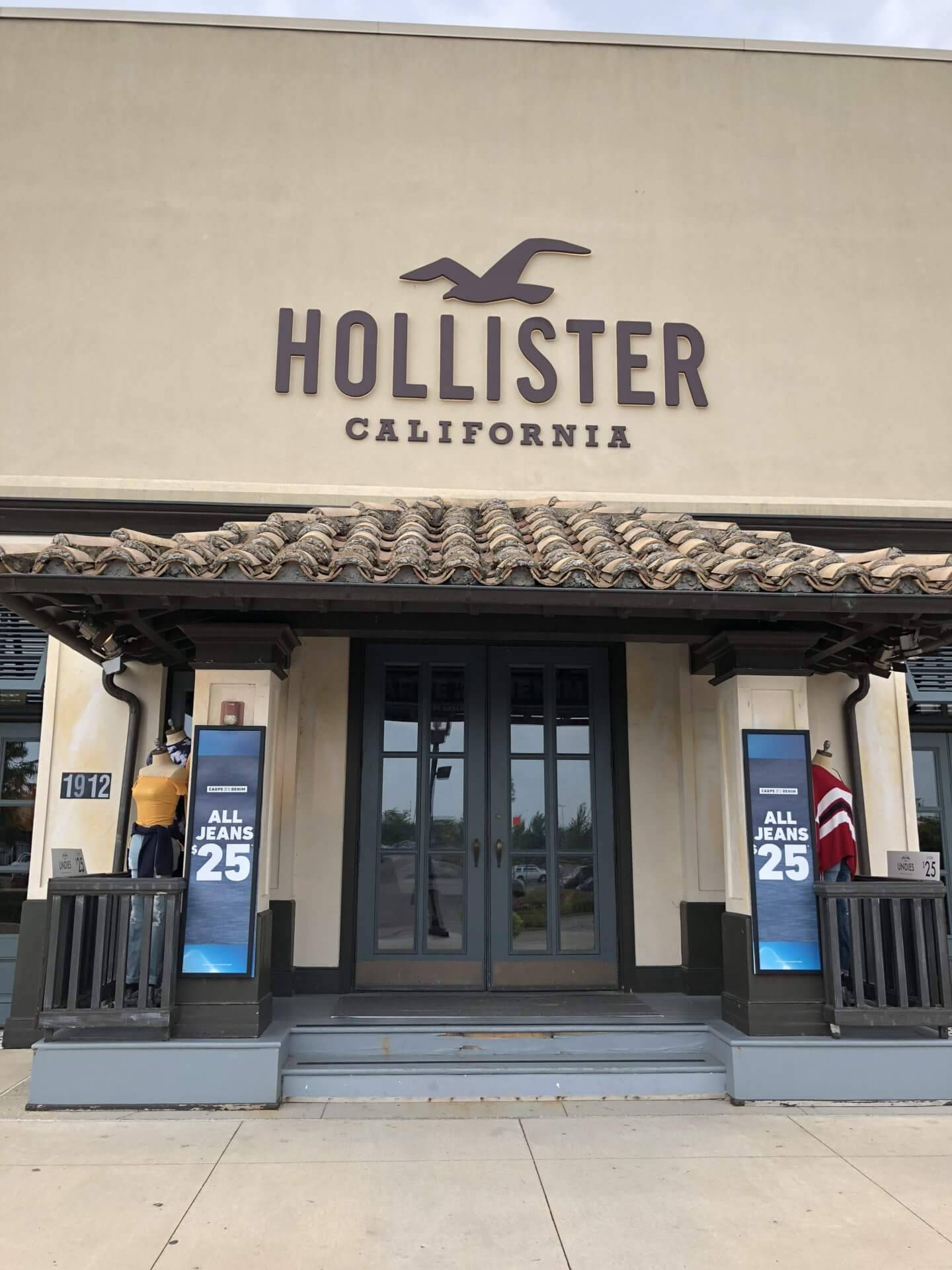 Hollister 店面標誌 壁紙 — Hollister California 商店的正面視圖，設有瓦片屋頂、人體模型和所有牛仔褲 25 減價標誌。