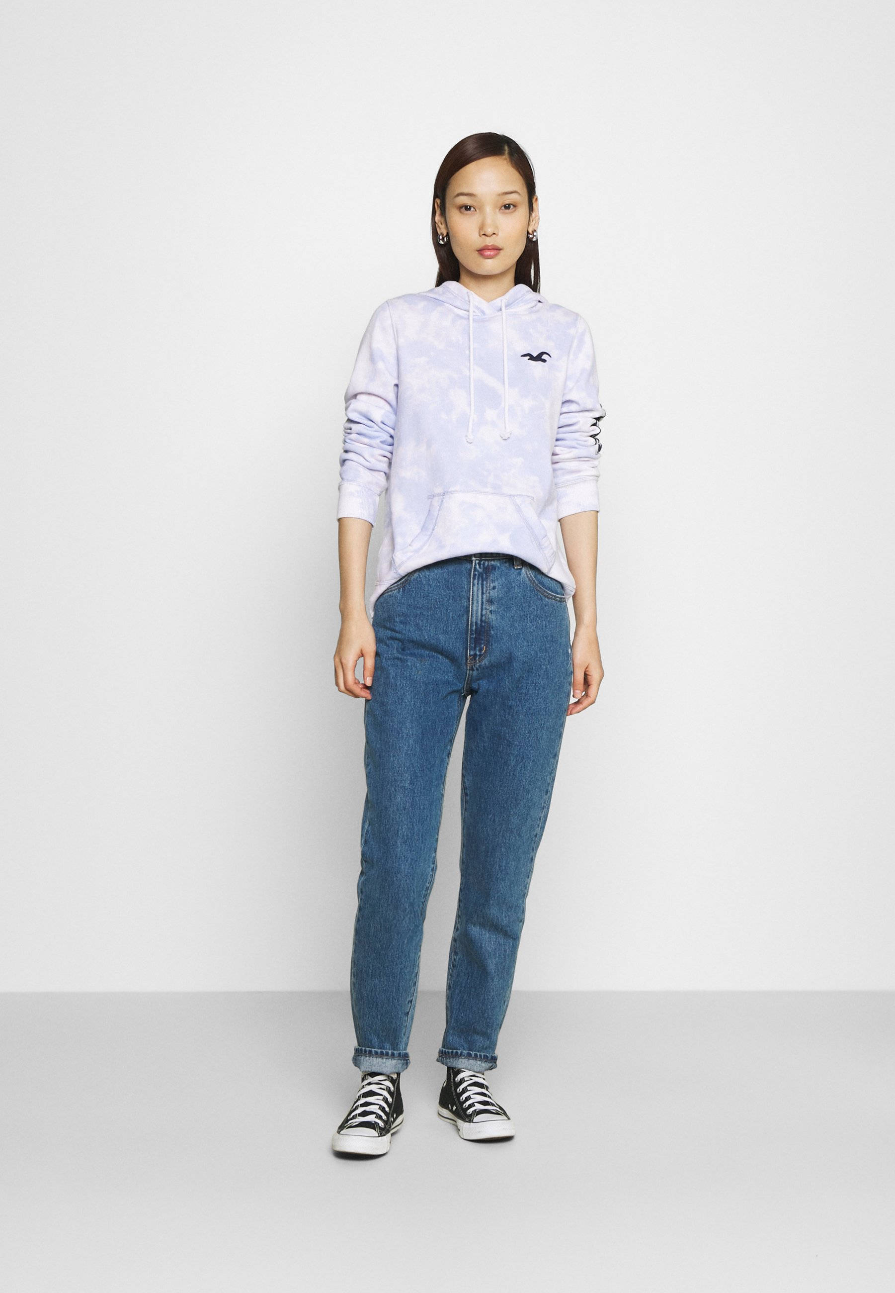 Hollister 淡白紫色連帽外套 壁紙 — 一位女士自信地站在純白色工作室背景前，身穿淺紫色紮染連帽衫、藍色牛仔褲和黑色運動鞋。