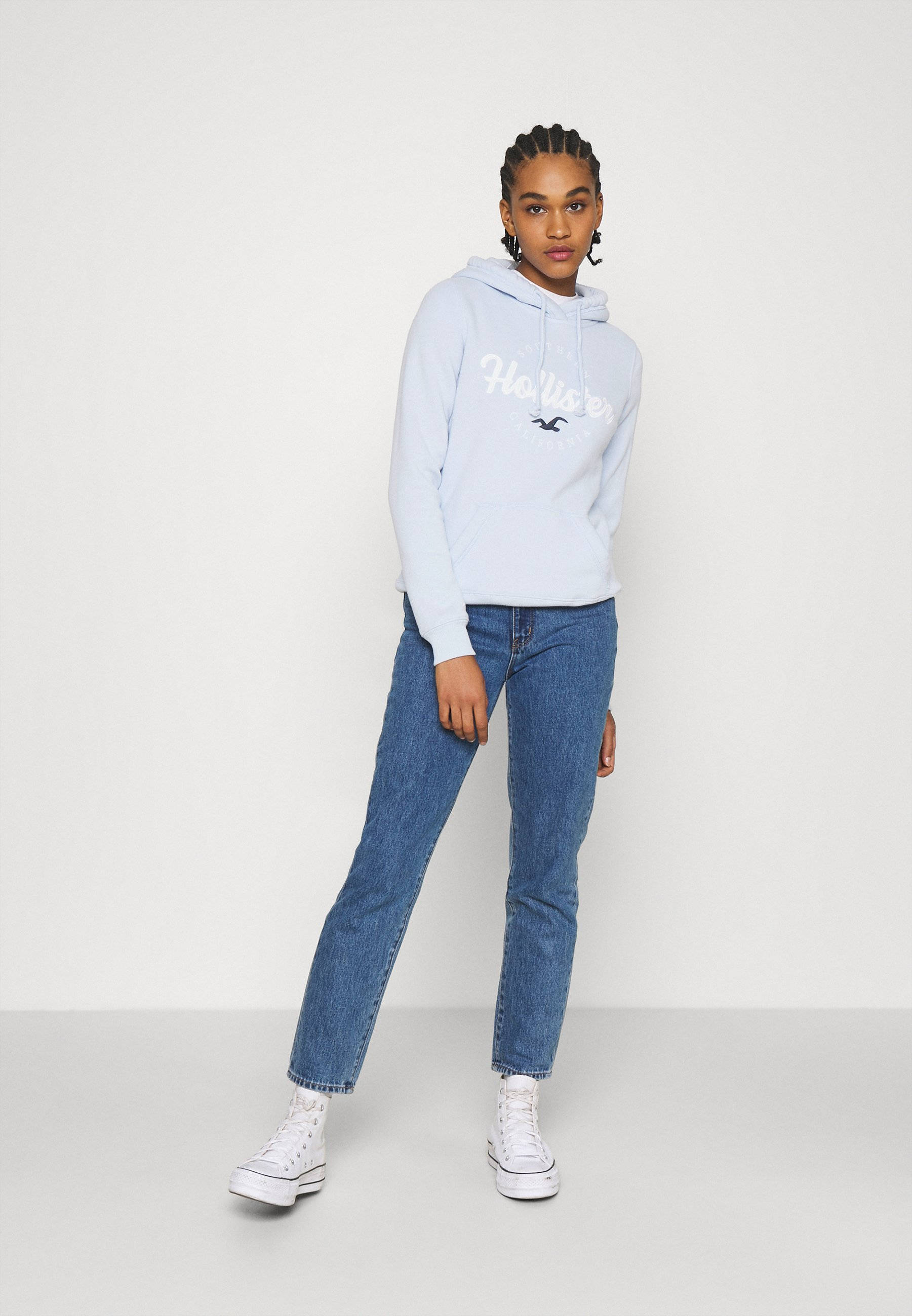 Hollister 淡藍色時尚連帽外套 壁紙 — 一位年輕女性身穿淺藍色Hollister連帽衫和藍色牛仔褲，自信地站在純白色攝影棚背景前。