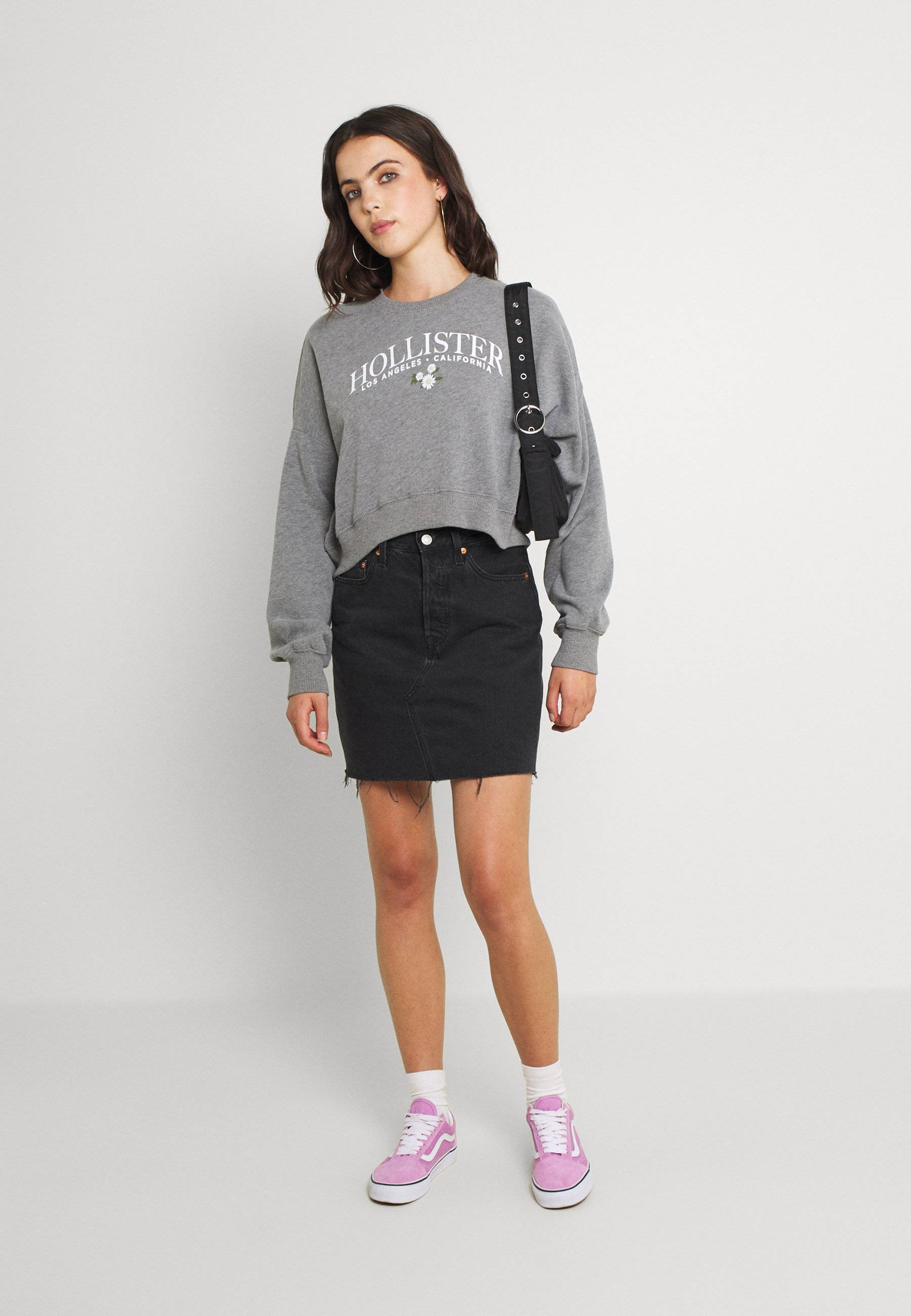 Hollister 深灰色衛衣 壁紙 — 一位年輕女性穿著灰色Hollister衛衣、黑色牛仔裙和粉紅色運動鞋，在純白色攝影棚背景前擺姿勢。
