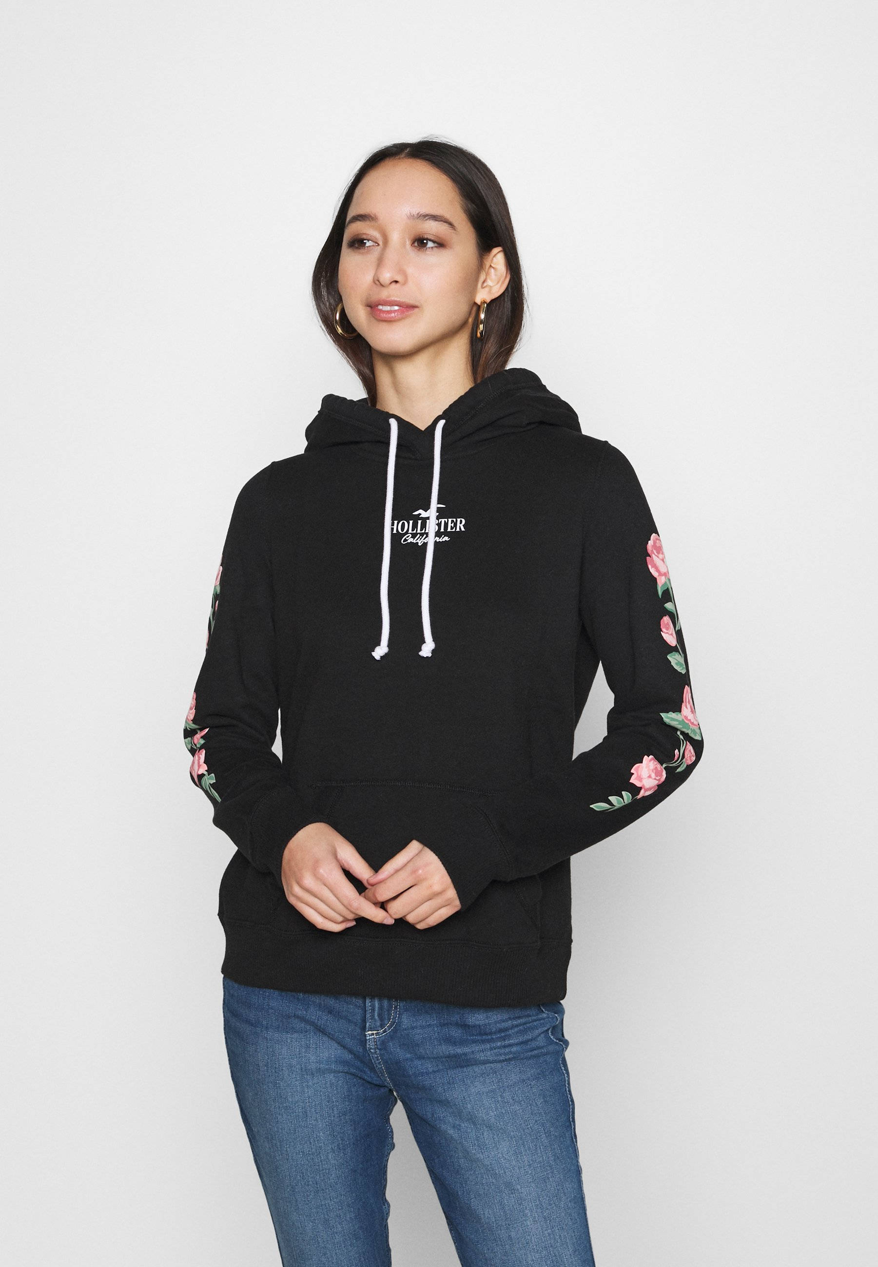 Hollister 粉紅玫瑰風格設計 壁紙 — 一名身穿黑色連帽衫、袖子上有粉紅色花卉刺繡的女性站在純白色背景前。