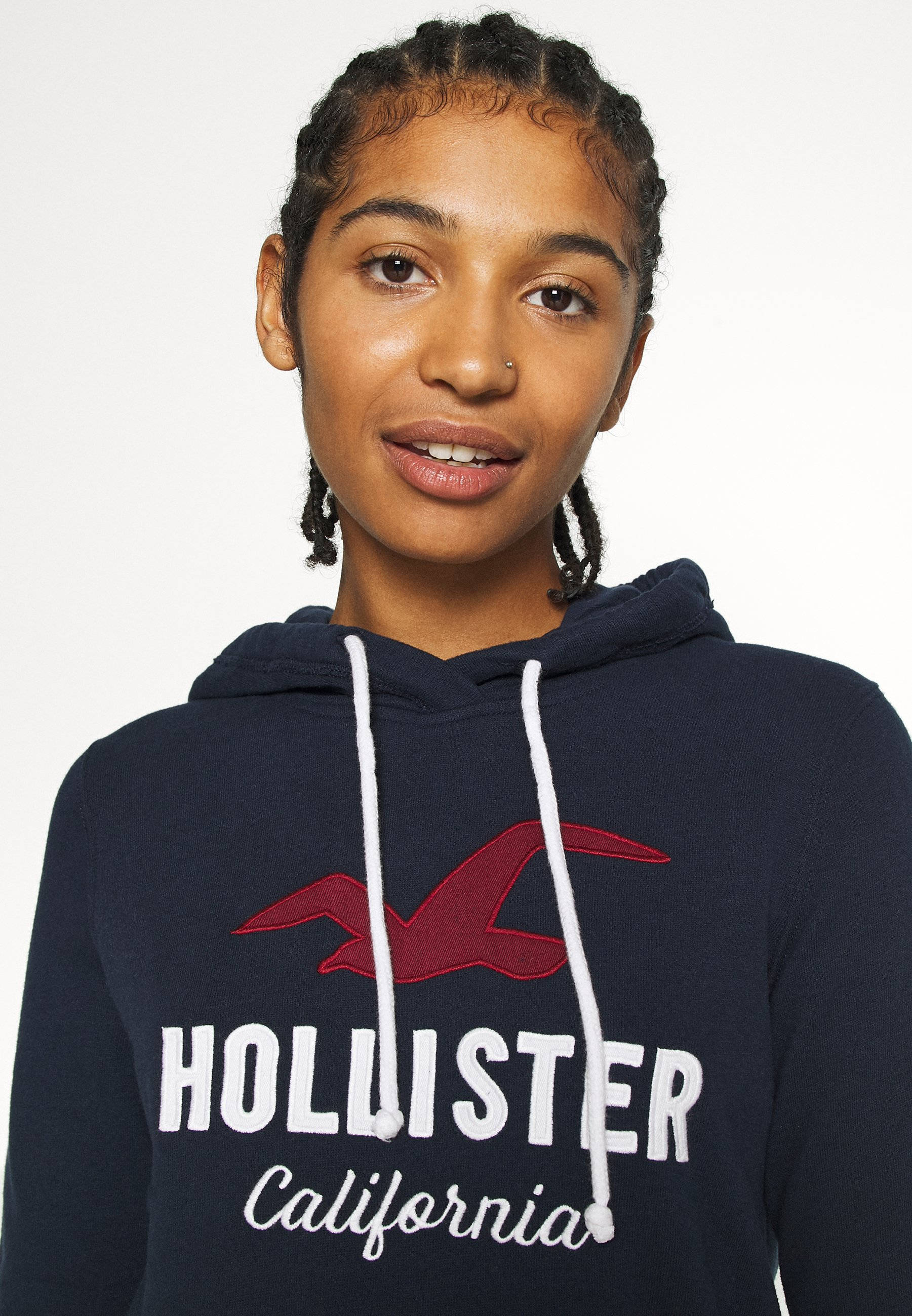 Hollister 紅色海鷗標誌 壁紙 — 一位辮子髮型的年輕女性，在純白色背景前穿著帶有紅色標誌和白色抽繩的深藍色Hollister連帽衫。