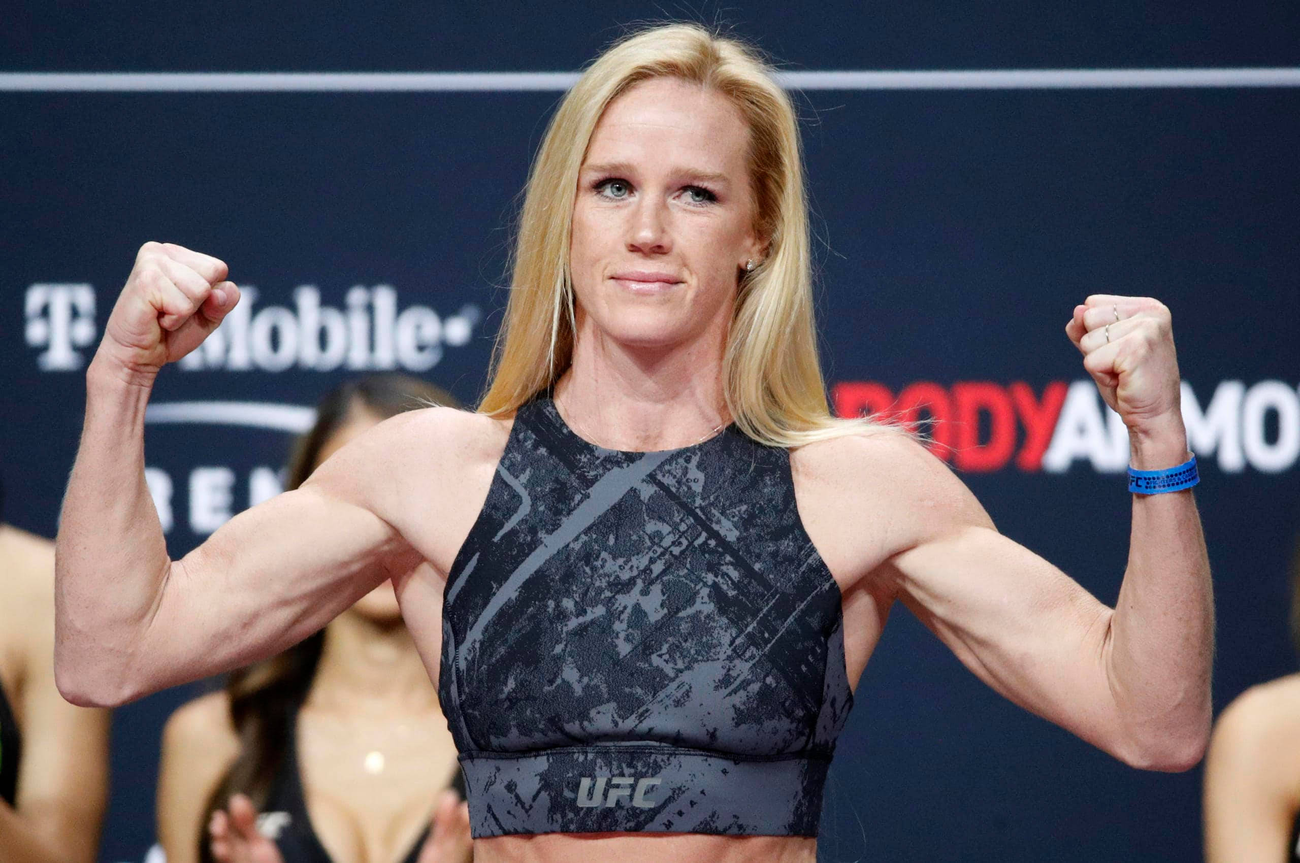 Holly Holm UFC體重確認儀式 壁紙 — 一位肌肉發達的金髮女性在品牌背景的稱重活動中，穿著UFC運動內衣彎曲雙臂。