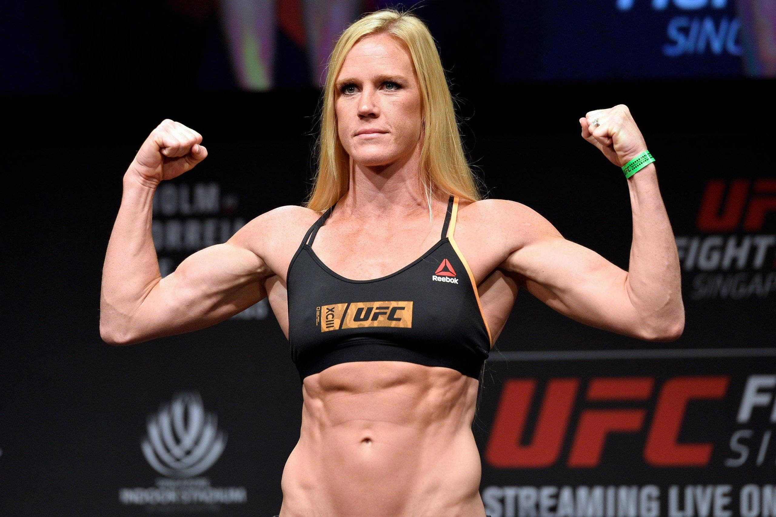 Holly Holm 練出的腹肌與肱二頭肌 壁紙 — 一位肌肉發達的女拳手在明亮的競技場燈光下，穿著黑色UFC運動內衣彎曲手臂。