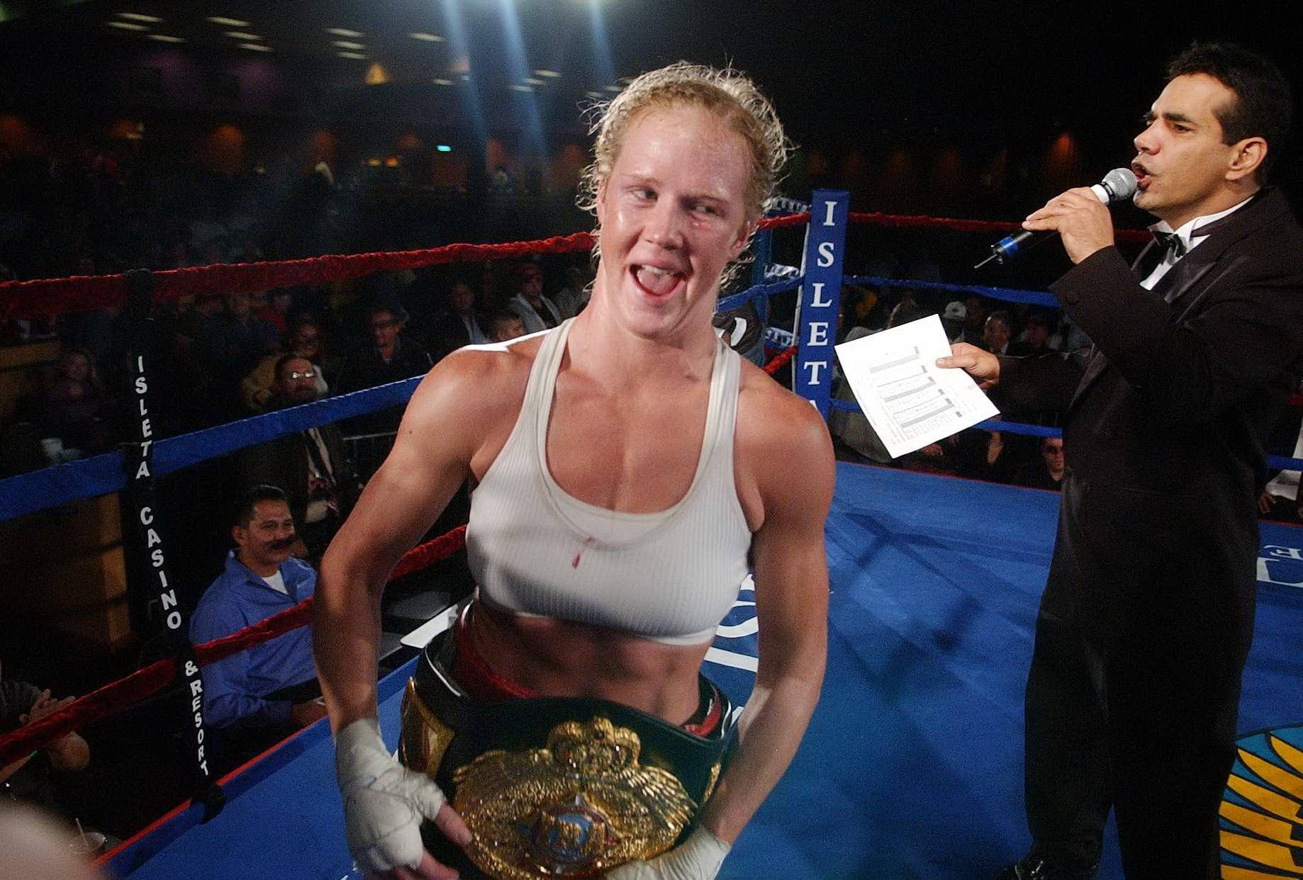 Holly Holm 舞動 IBA世界拳王腰帶 壁紙 — 一位身穿白色背心和冠軍腰帶的勝利女拳擊手在燈光明亮的拳擊場中咧嘴大笑，旁邊一位解說員正對著麥克風說話。