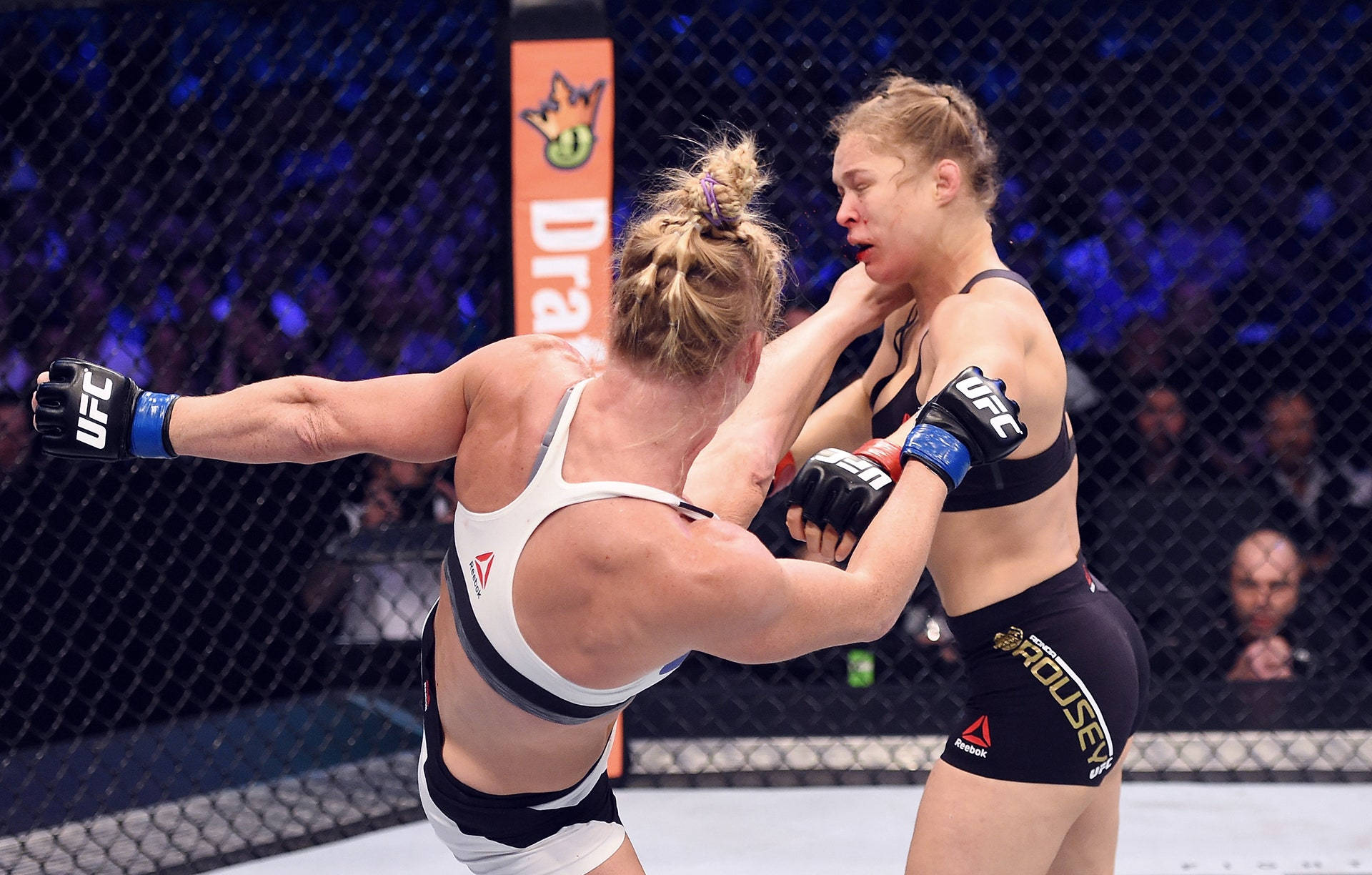 Holly Holm 踢中 Ronda Rousey 臉部 壁紙 — 兩名女性MMA拳手在明亮的場館燈光下，於UFC八角籠內激烈纏鬥。