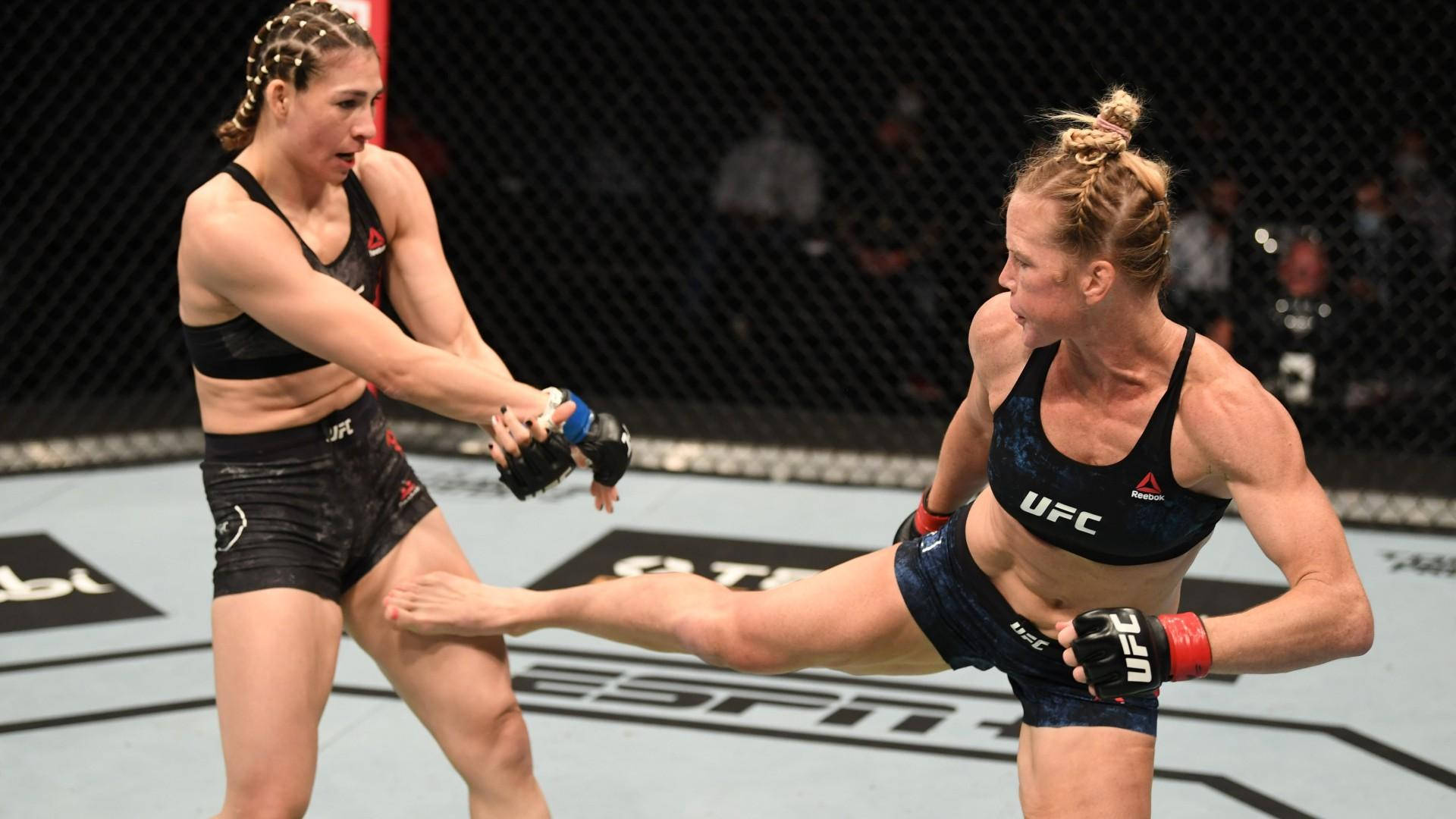 Holly Holm 踢擊墨西哥新星 Irene Aldana 壁紙 — 兩名女性MMA拳手在籠中交鋒，一人使出高踢，另一人阻擋。