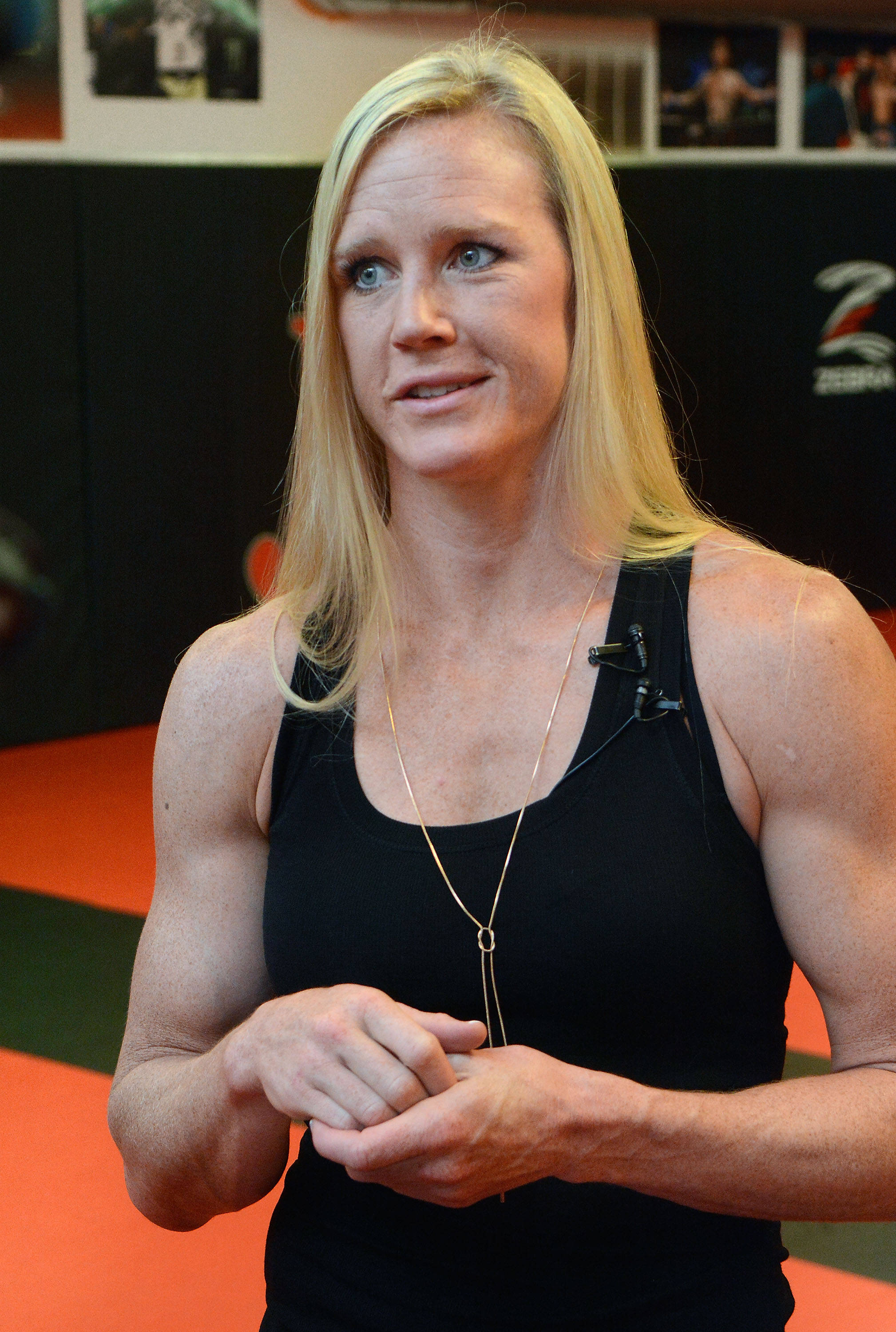 Holly Holm 金色長髮 壁紙 — 一位肌肉發達的金髮女性穿著黑色背心，自信地站在一個有橙色墊子和拳擊海報的健身房裡。