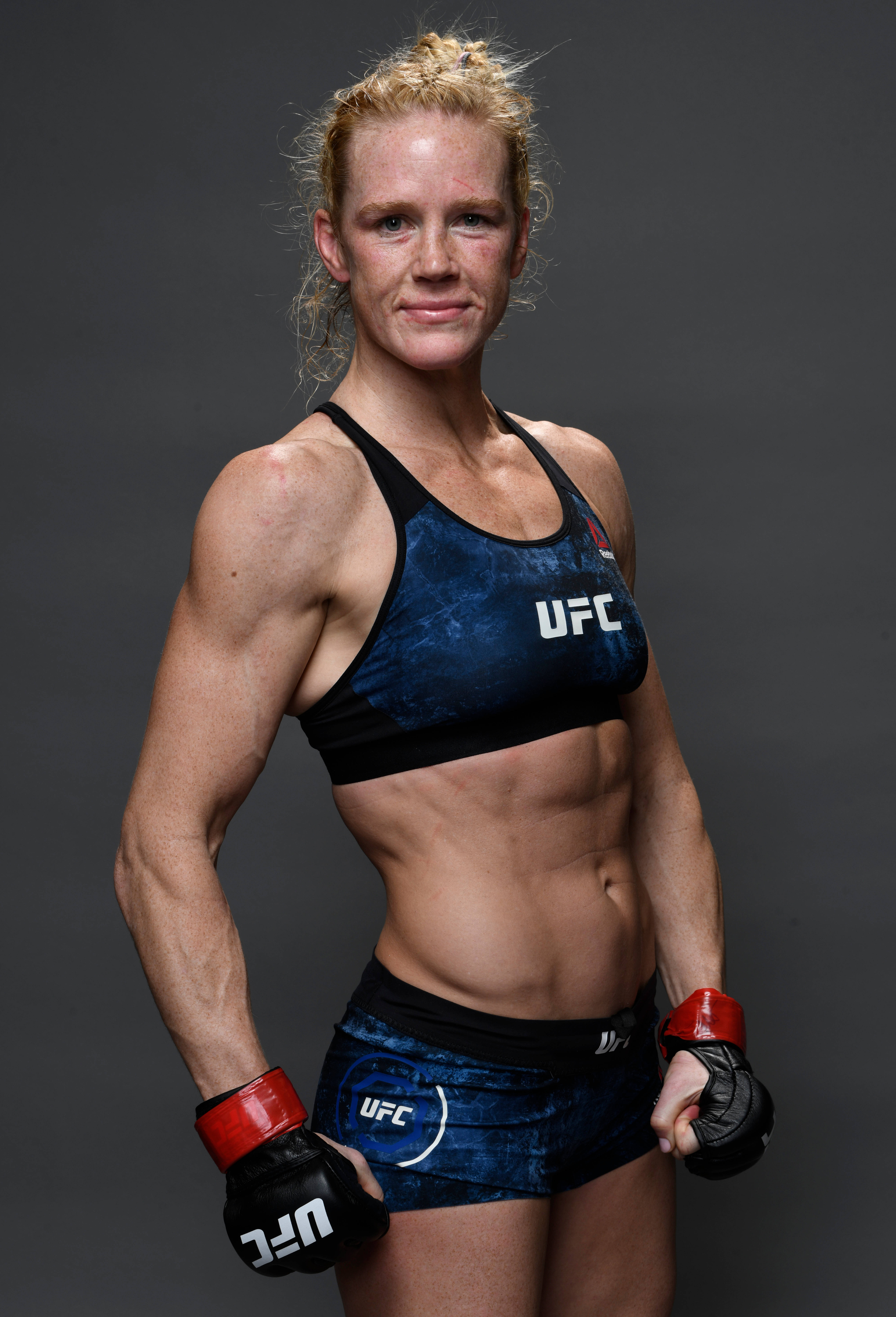 Holly Holm展現健美身材 壁紙 — 一位肌肉發達的女性UFC拳手，身穿藍色裝備和紅色手套，在深灰色工作室背景前自信地擺姿勢。