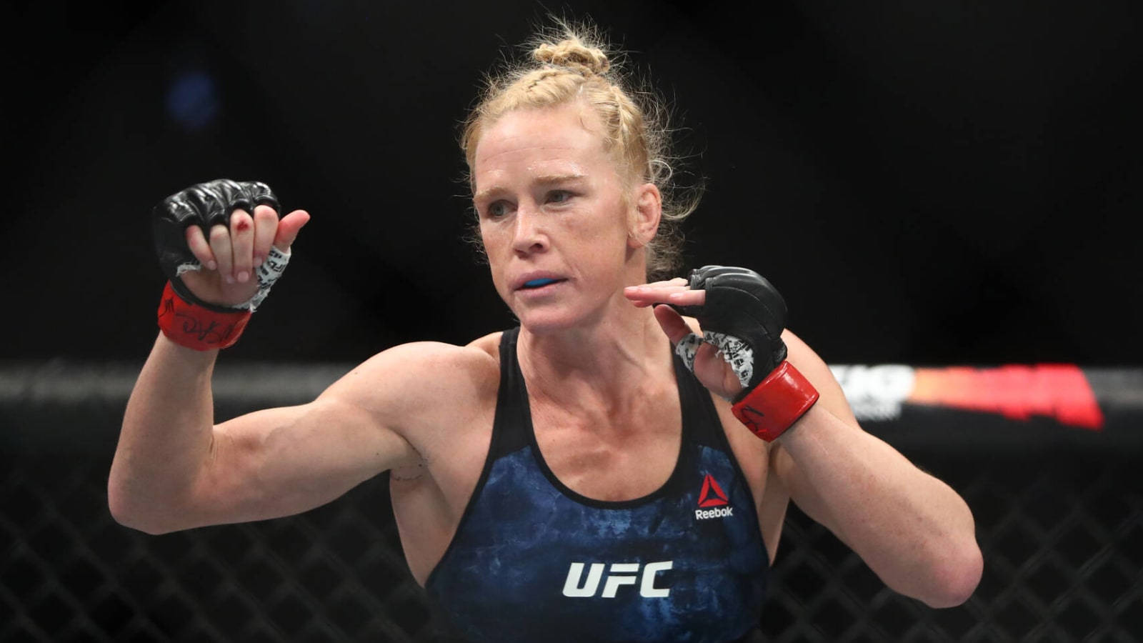 Holly Holm的MMA戰鬥姿勢 壁紙 — 一位專注的女性MMA拳擊手，身穿藍色UFC背心和紅色手套，在黑暗的八角籠中準備就緒。