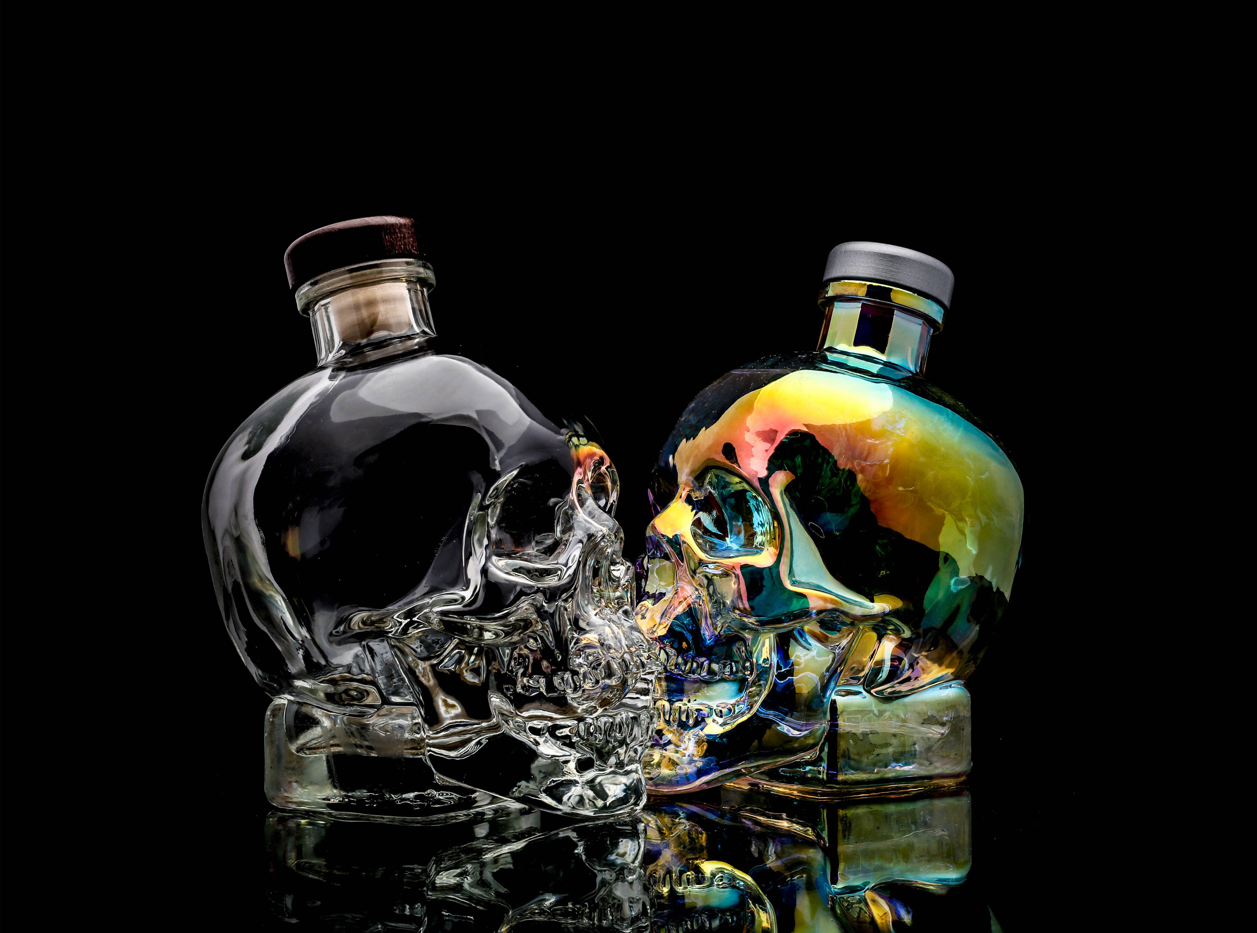 Holographic Crystal Head伏特加 壁紙 — 兩個骷髏形狀的酒瓶，一個透明，一個彩虹色，映照在光亮的黑色表面上。