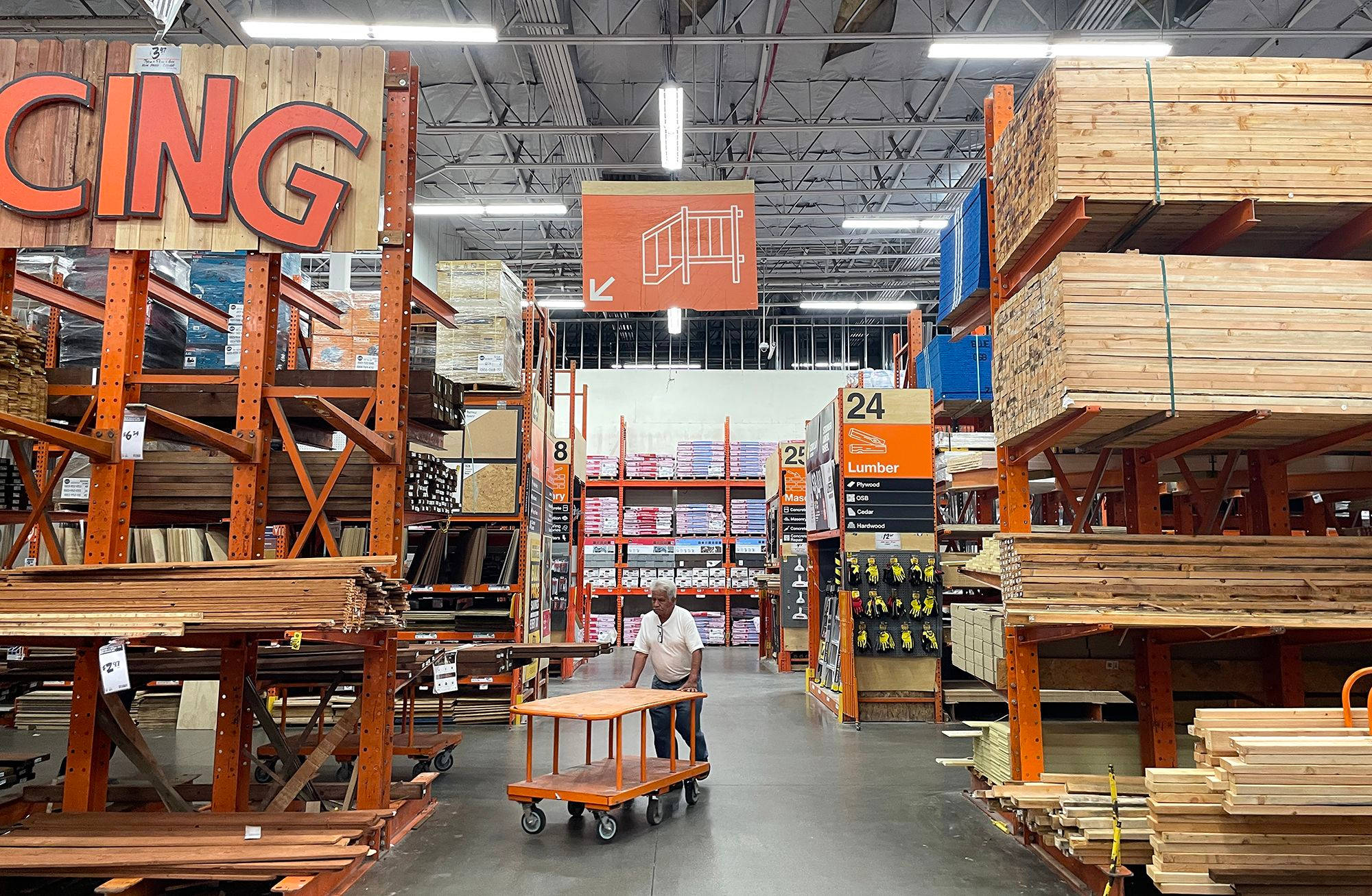 Home Depot 木料區 壁紙 — 一名男子推著手推車，穿過寬敞的五金店貨架通道，兩旁堆滿高聳的木材和建築用品，頭頂是明亮的螢光燈。