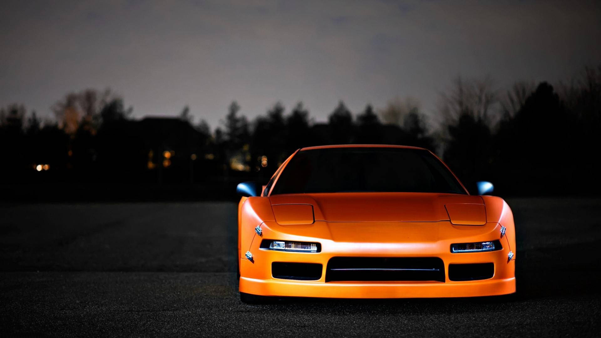 橙色Honda NSX JDM 壁紙 — 一輛充滿活力的橙色跑車在暮色中閃耀，車頭燈照亮黑暗的瀝青道路和樹影。