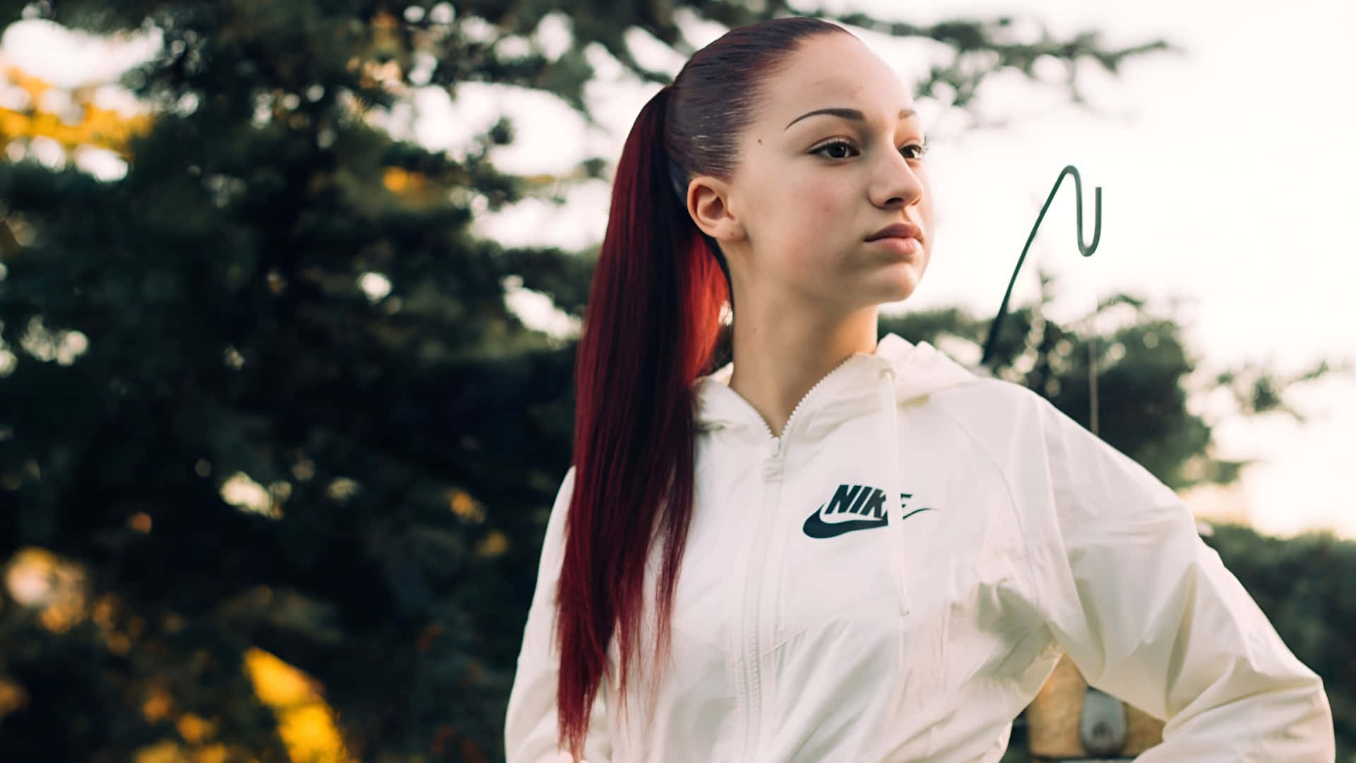  壁紙 — 一位年輕女性紮著充滿活力的紅色馬尾辮，穿著白色Nike風衣，背景是柔和模糊的樹木。