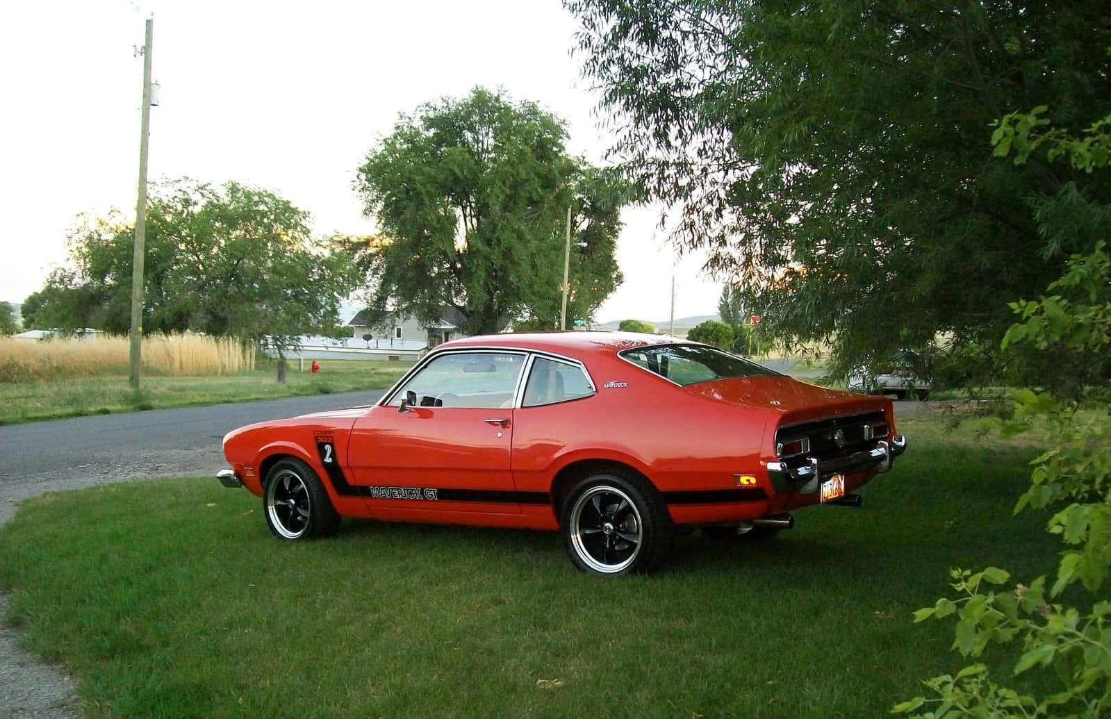 Ford Maverick 桌布 壁紙 — 一輛鮮紅色經典肌肉車，帶有黑色條紋，停在鄉村道路旁的草地上，背景是柔和的傍晚天空。