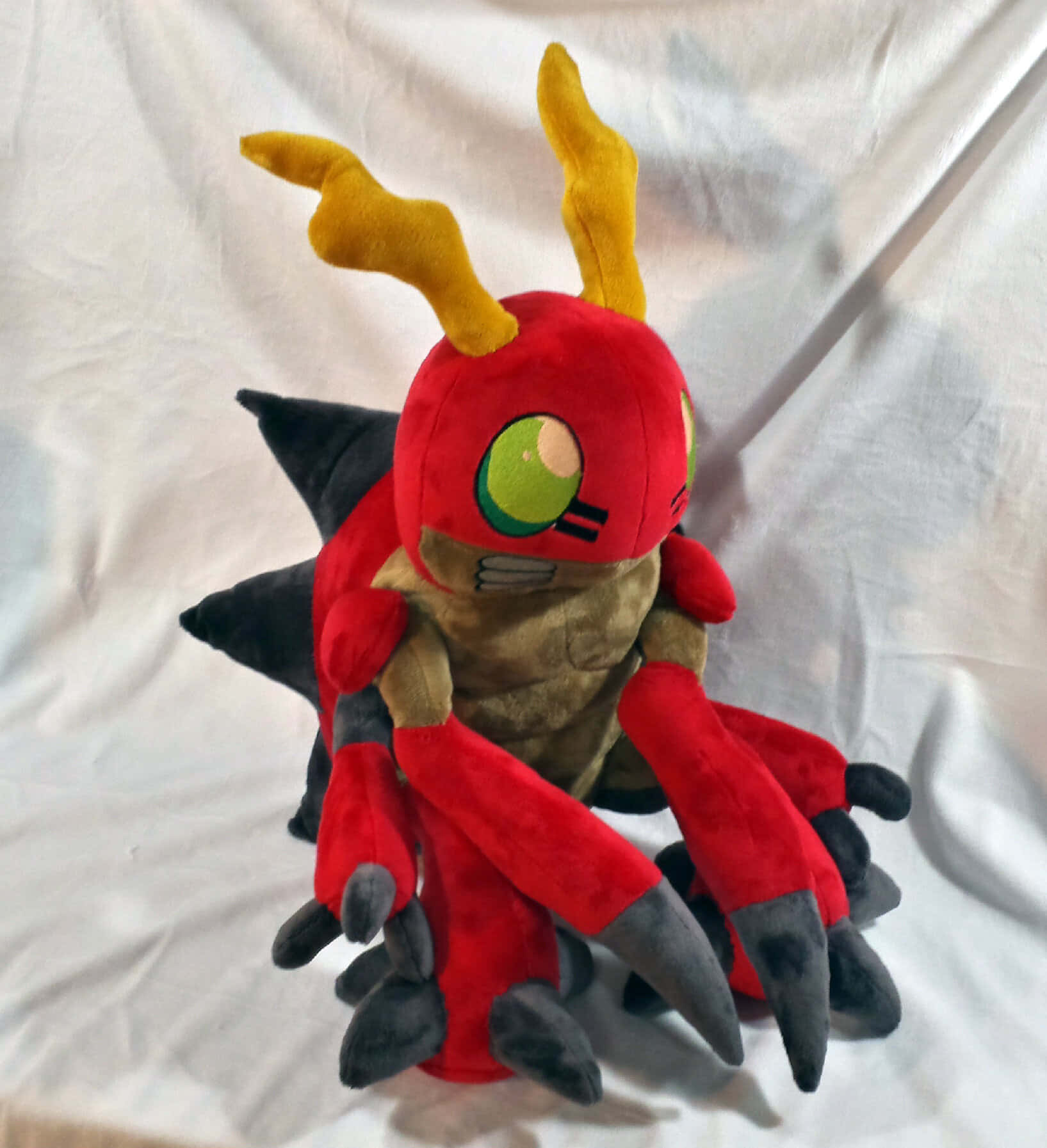 Digimon Tentomon 桌布 壁紙 — 一個紅色毛絨玩具，有黃色角、綠色眼睛和黑色爪子，坐在白色布料背景上。