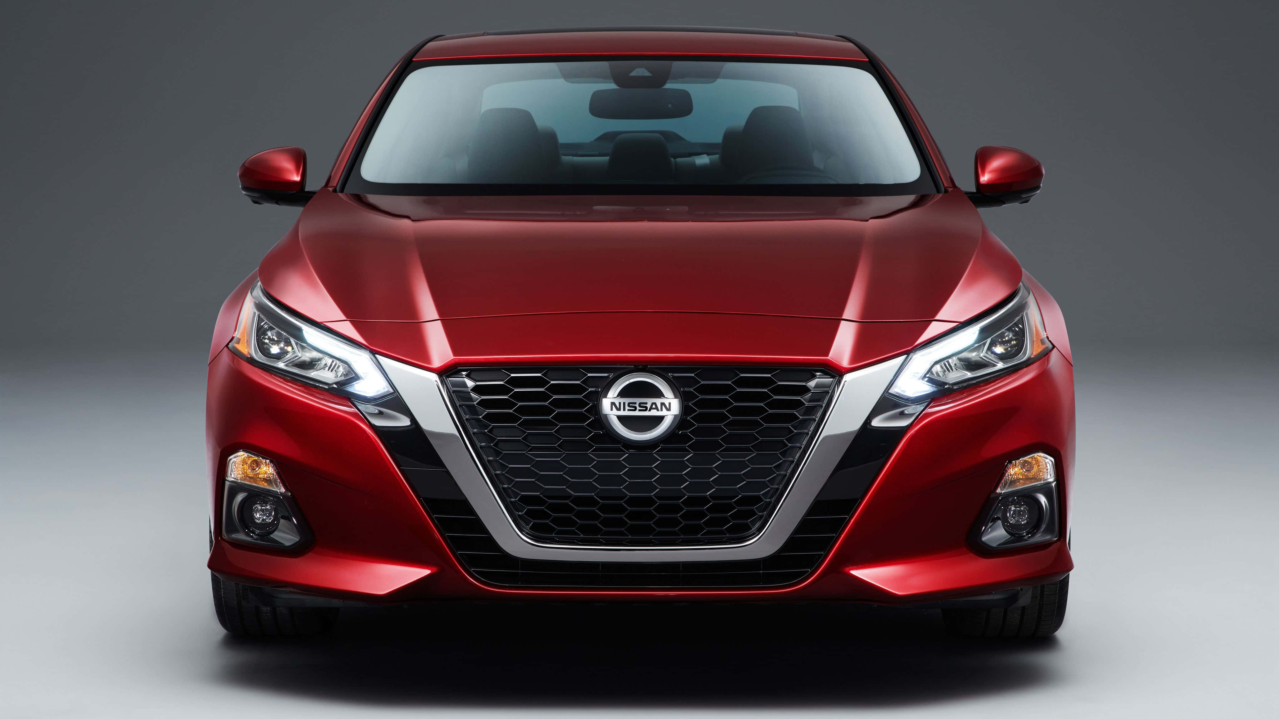 Nissan Altima 桌布 壁紙 — 一輛光亮的紅色日產房車正面朝前，在攝影棚內以戲劇性燈光突顯其時尚現代的水箱護罩和銳利頭燈。