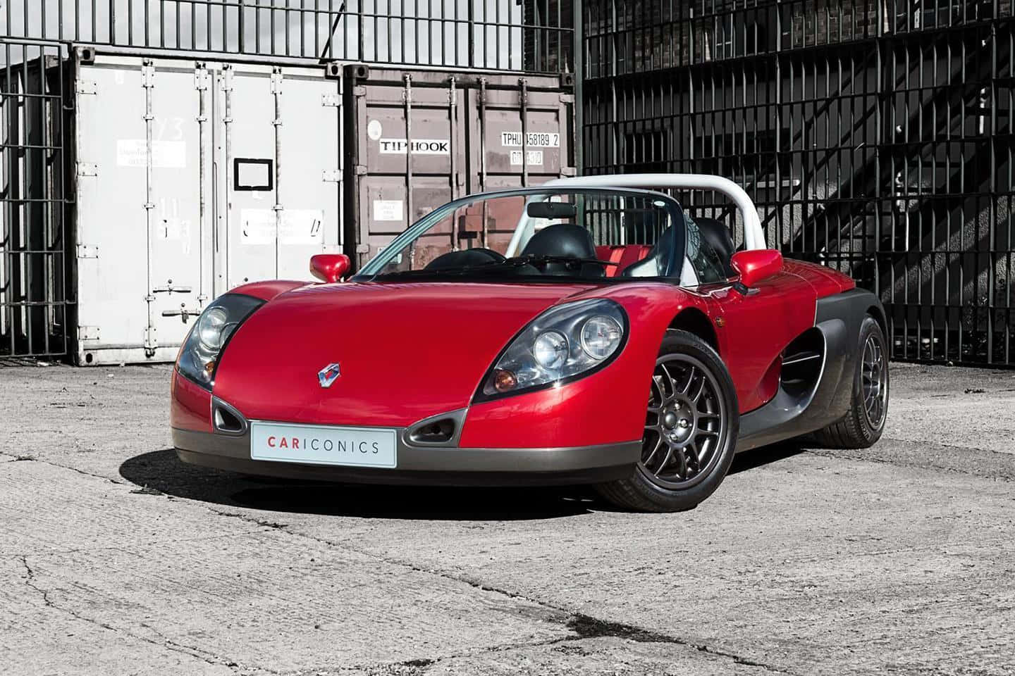 Renault Sport Spider 桌布 壁紙 — 一輛充滿活力的紅色跑車，帶有黑色飾邊和白色防滾架，停在裂開的混凝土上，前面是貨運集裝箱和黑色金屬圍欄。