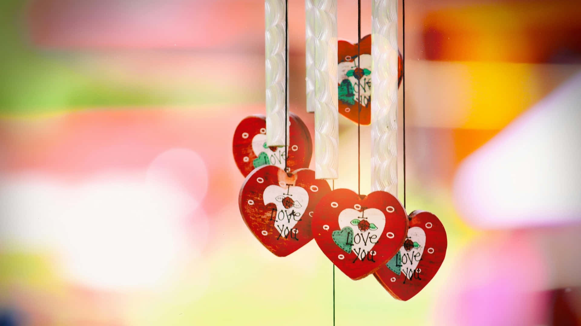  壁紙 — 紅色心形風鈴，上面刻有「Love You」字樣，懸掛在柔和模糊的彩虹背景前。