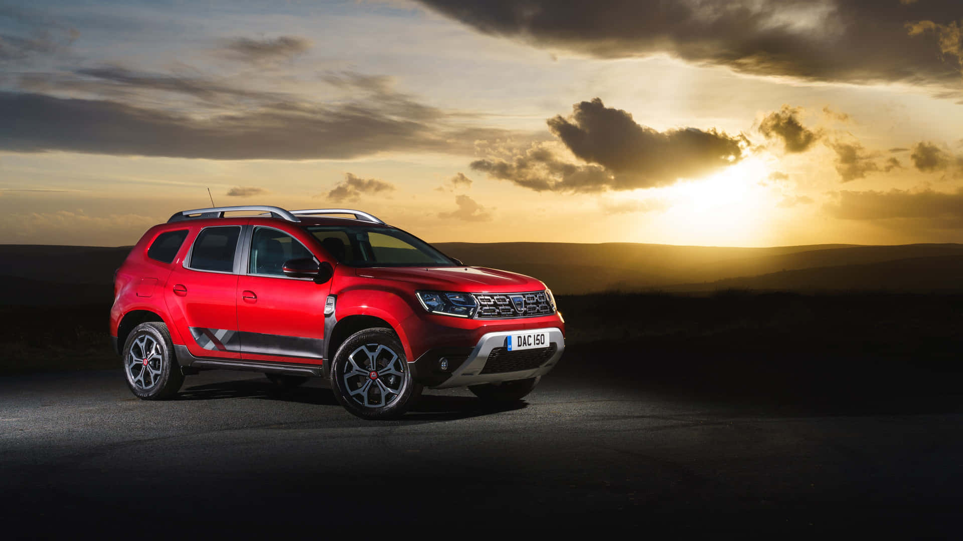  壁紙 — 一輛鮮紅色的Dacia Duster SUV停在黑暗的道路上，金色的夕陽光線穿過戲劇性的雲層，灑在起伏的山丘上。