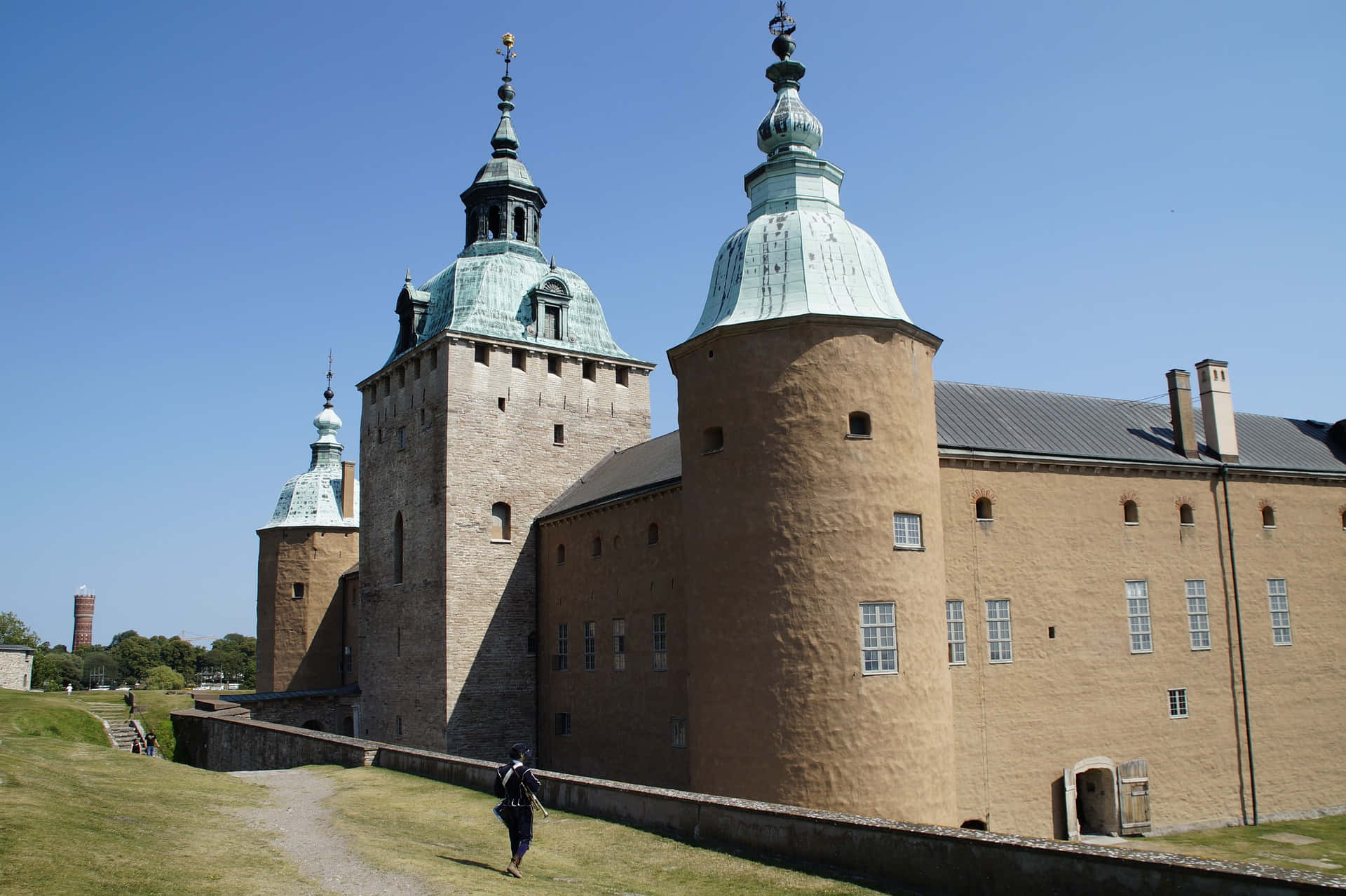 Kalmar Sweden 桌布 壁紙 — 一座宏偉的歷史城堡，擁有銅製圓頂塔樓，矗立在清澈的藍天下，一位孤獨的訪客沿著草地小徑行走。