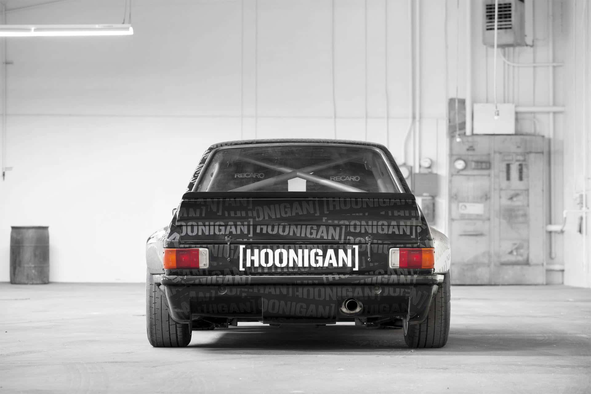 Hoonigan汽車工廠 壁紙 — 從後方觀看一輛黑色Hoonigan改裝車停放在單色調的簡陋工業車庫中。