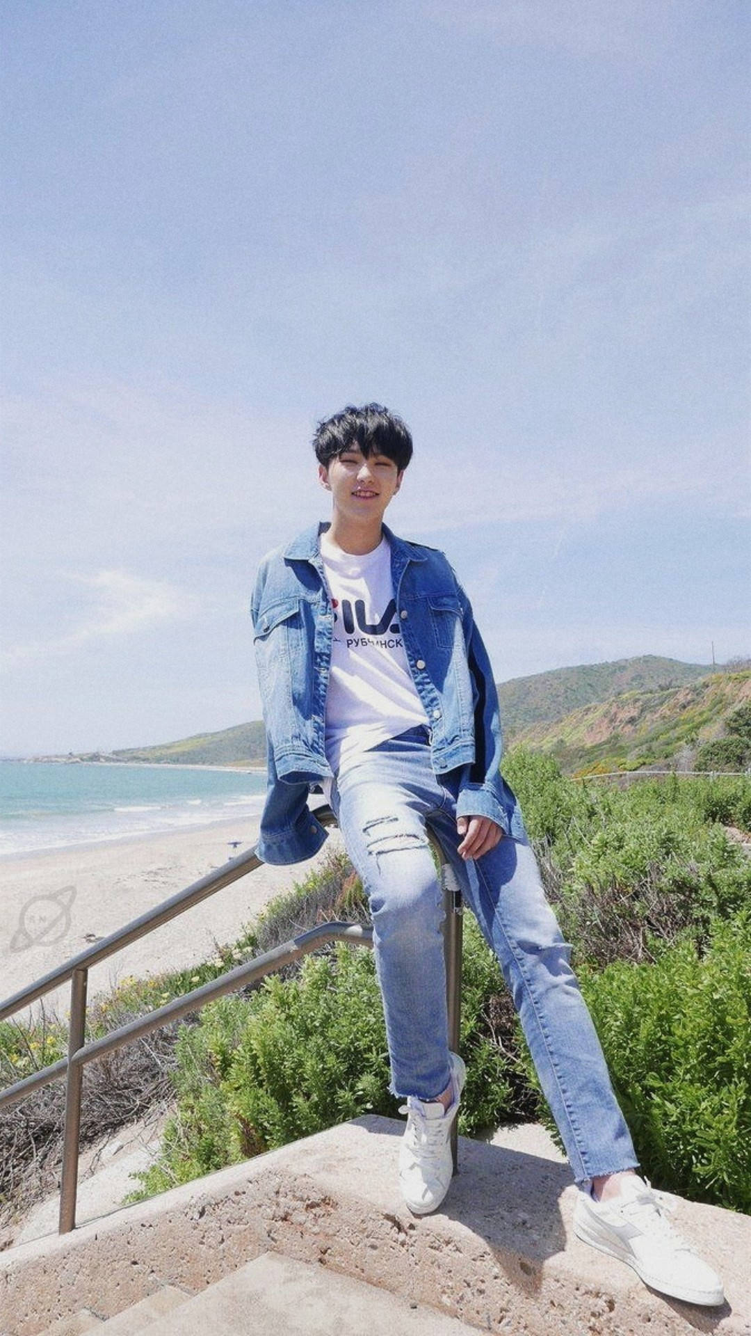 Hoshi 在濟州島 壁紙 — 一名身穿牛仔外套和破洞牛仔褲的年輕男子，隨意地坐在石階上，俯瞰著陽光普照的海灘，碧綠的海水和翠綠的山丘，天空清澈湛藍。