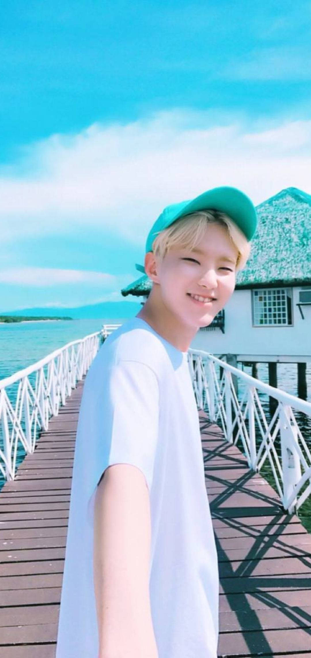 Hoshi 海邊風景 壁紙 — 一個戴著綠色帽子的微笑年輕人在木製碼頭上俯瞰著碧綠色的海水，天空湛藍。