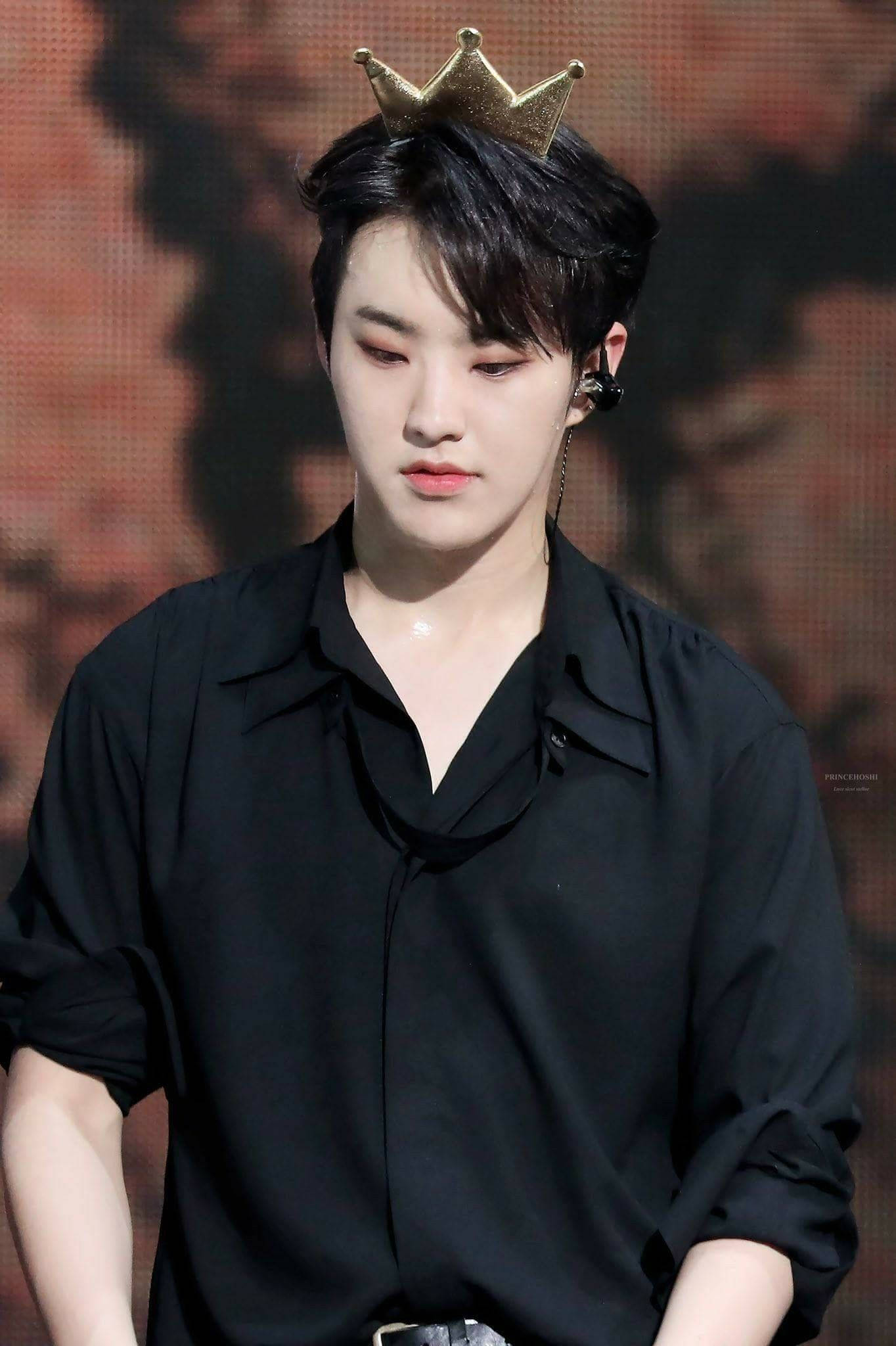Hoshi 黑色造型 壁紙 — 一位黑髮年輕男子戴著小金皇冠和黑色襯衫，背景是模糊的紅色舞台。