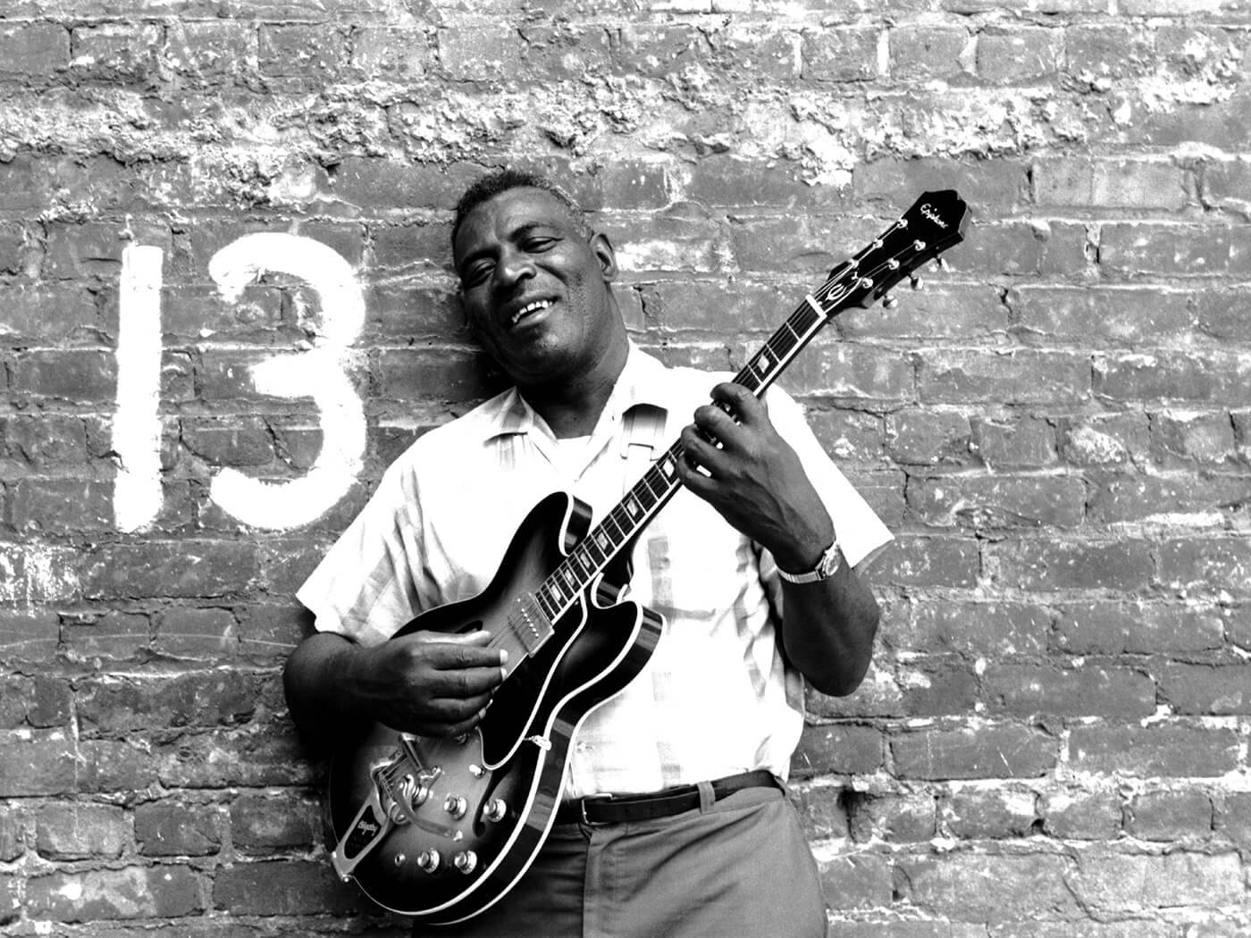 Howlin' Wolf《13號磚牆》黑白照片 壁紙 — 一位快樂的黑人男子微笑著，手持結他，背景是風化的磚牆，牆上漆有白色的數字13。