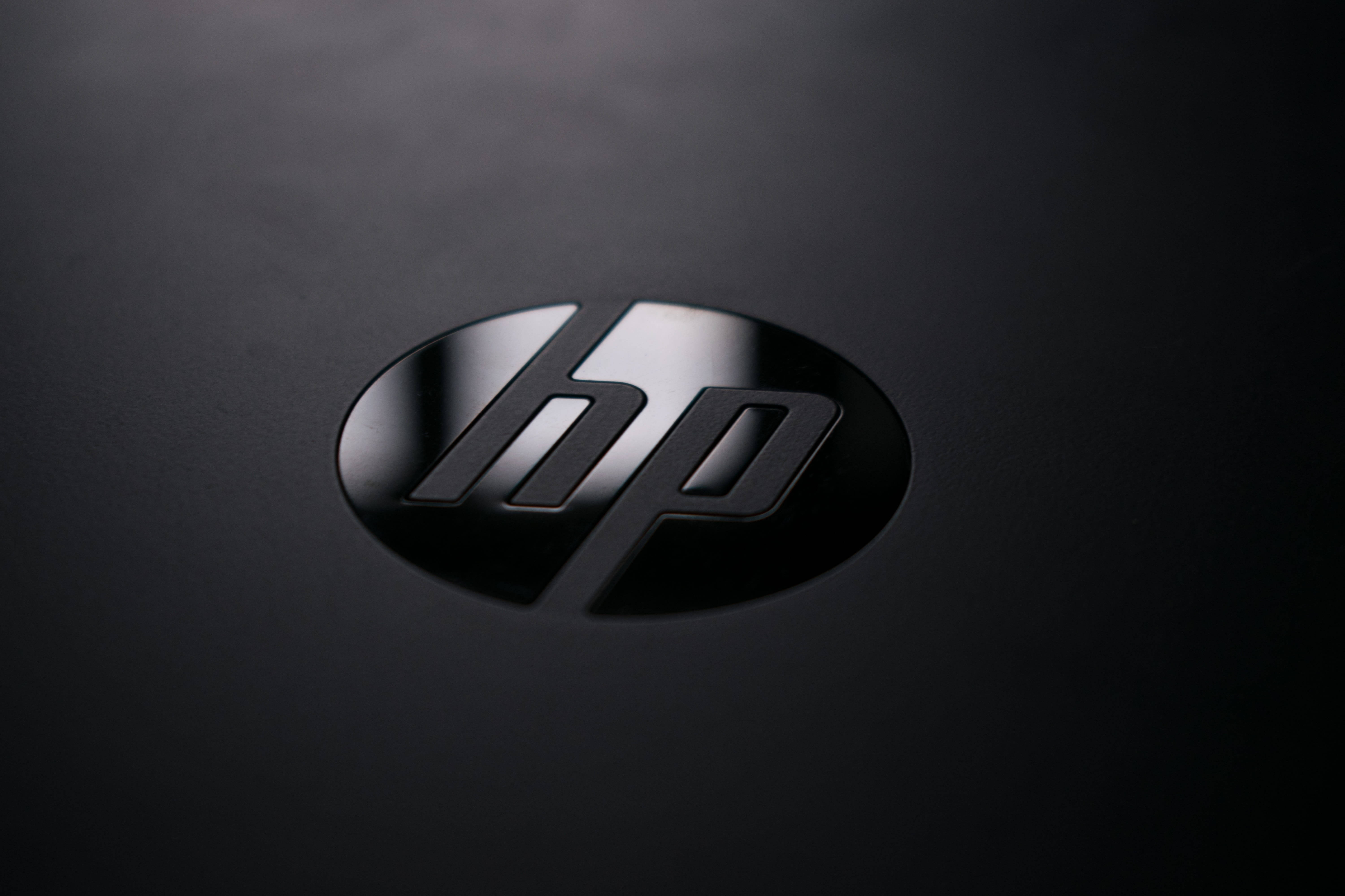 標語：時尚強勁的HP筆記本電腦 壁紙 — HP 標誌特寫，位於啞光黑色表面上，柔和光線突顯其金屬光澤。