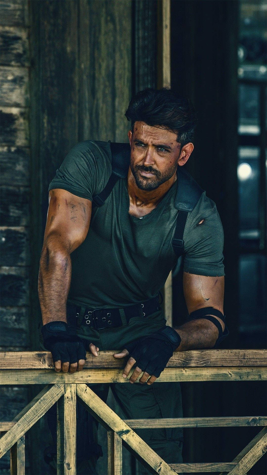 Hrithik Roshan《戰爭》電影劇照 壁紙 — 一個肌肉發達的男人穿著深色裝備，在昏暗的鄉村環境中，表情嚴肅地靠在木欄杆上。