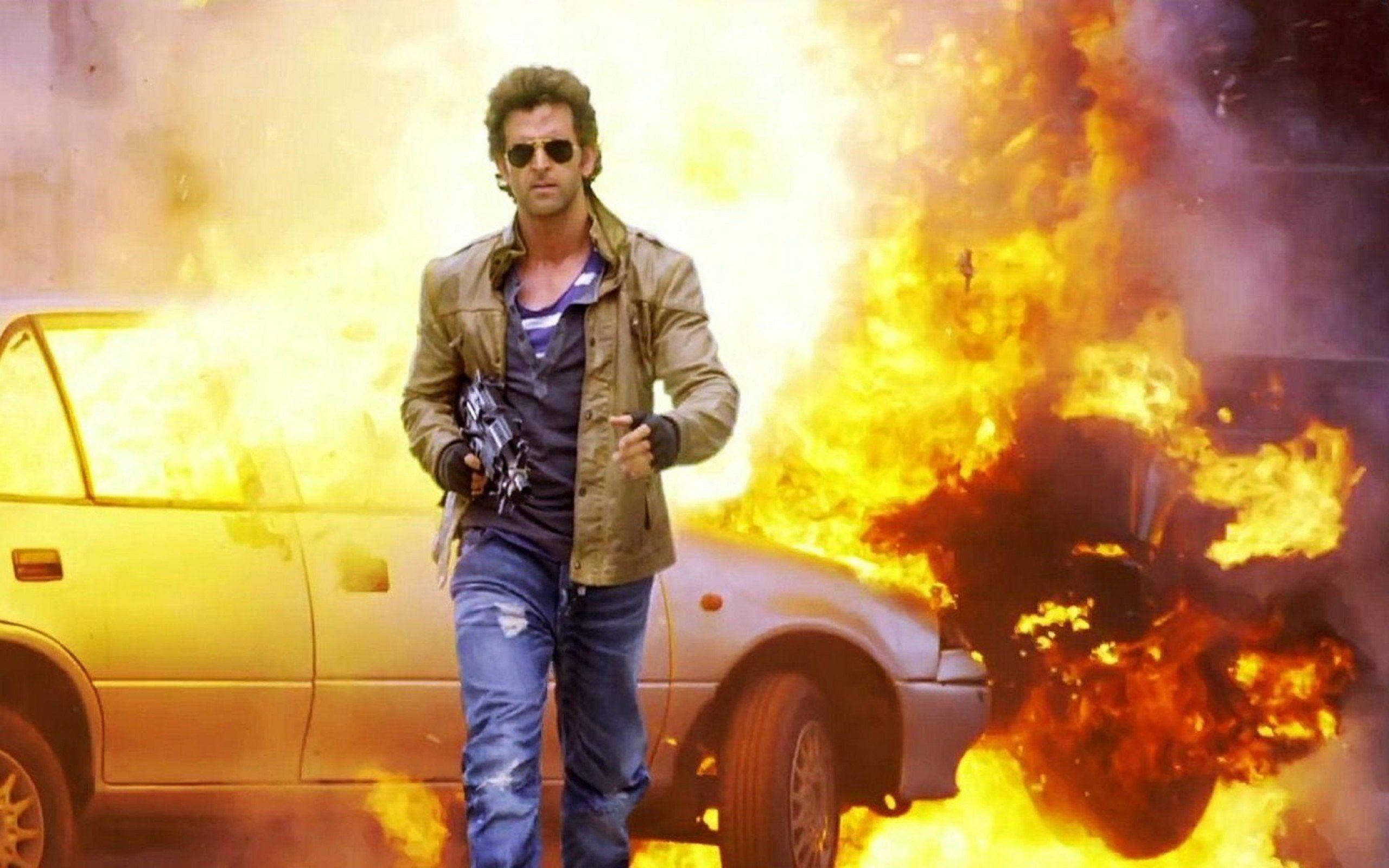 Hrithik Roshan 車輛爆炸劇照 壁紙 — 一個戴著太陽眼鏡和外套的男人挑釁地站在一輛被明亮橙色火焰吞噬的燃燒汽車旁邊。